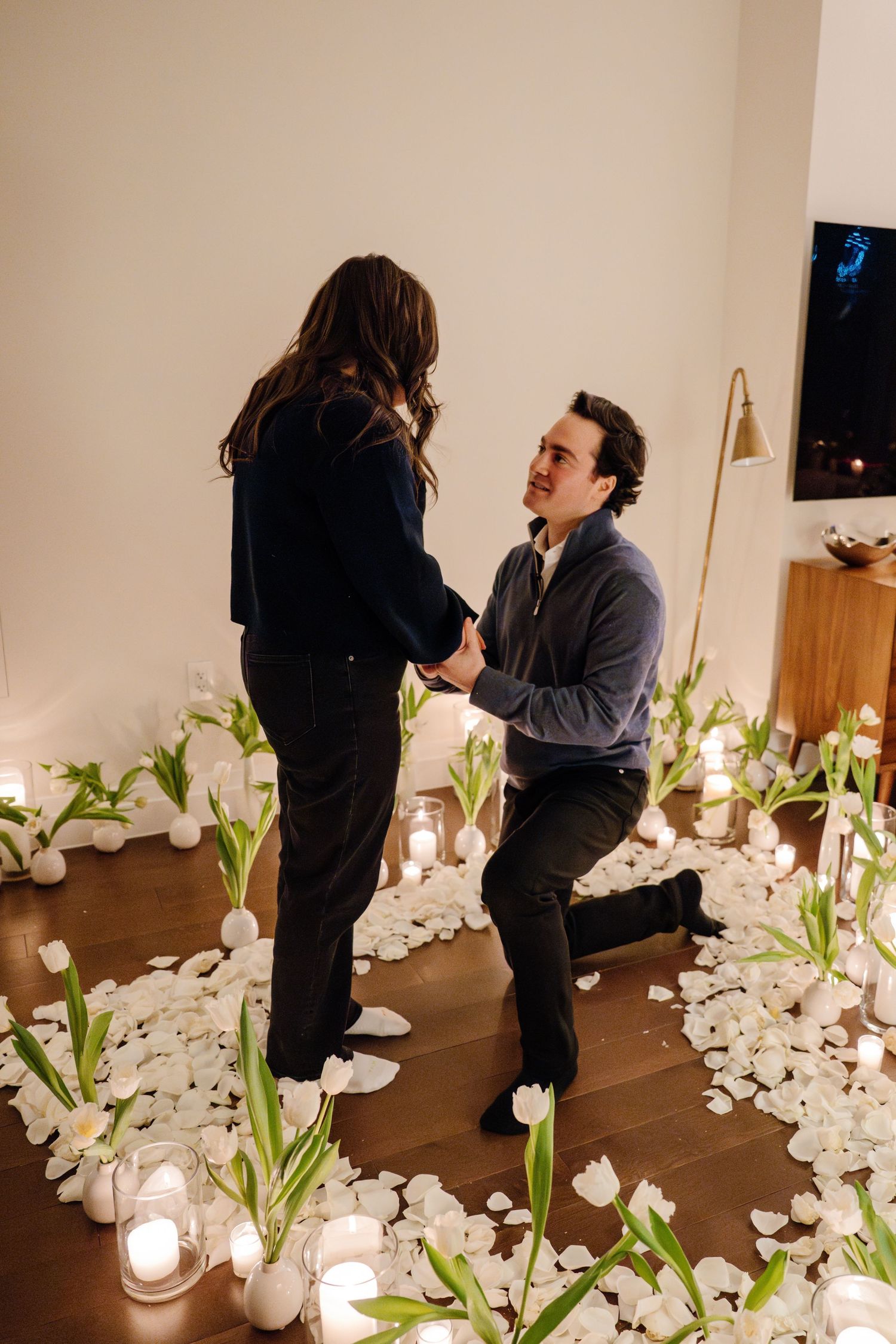 Man on one knee proposing to his girlfriend surrounded by white tulips, rose petals, and candles in an apartment.