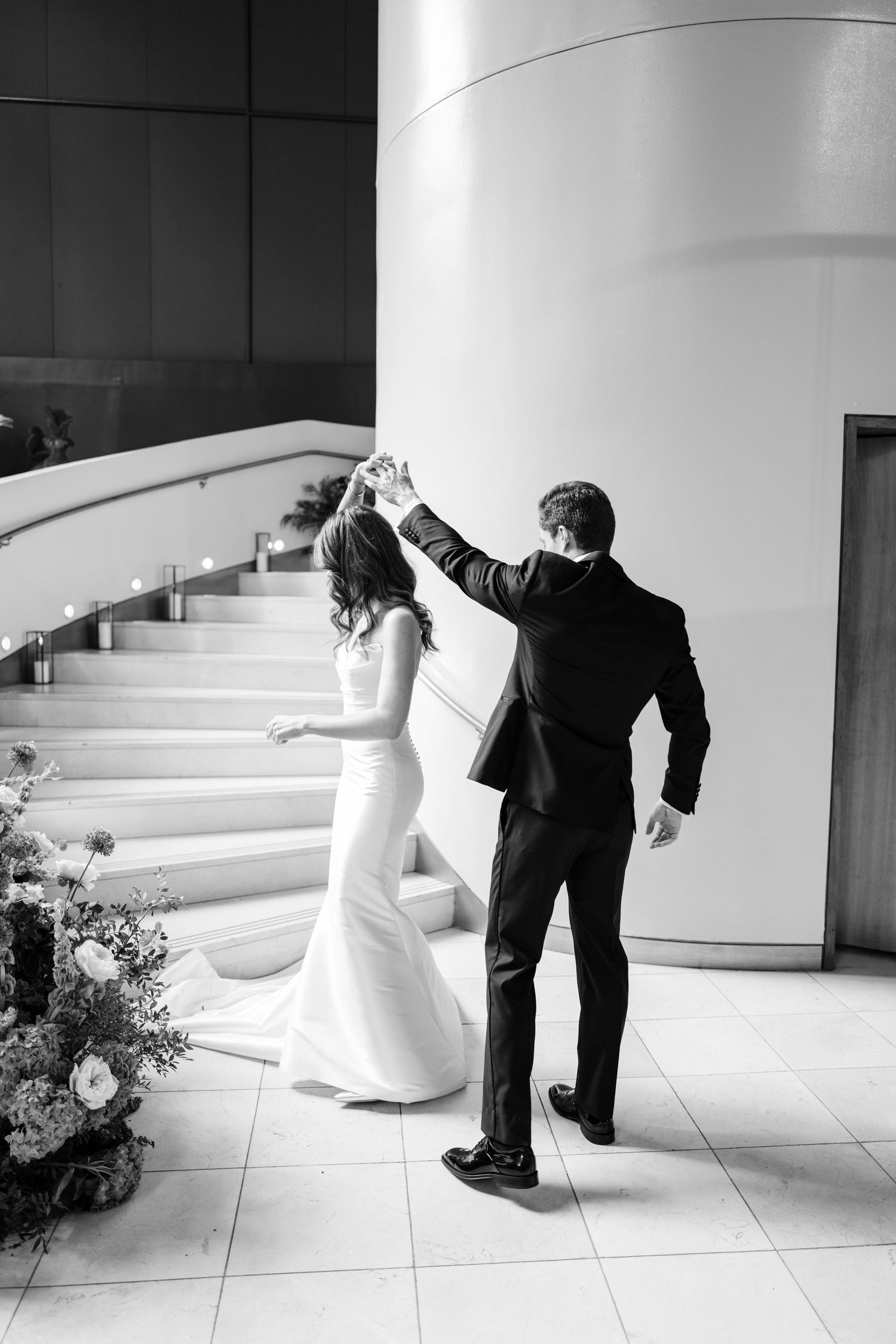 Black and white photo of groom twirling bride in wedding gown near floral arrangement at staircase.
