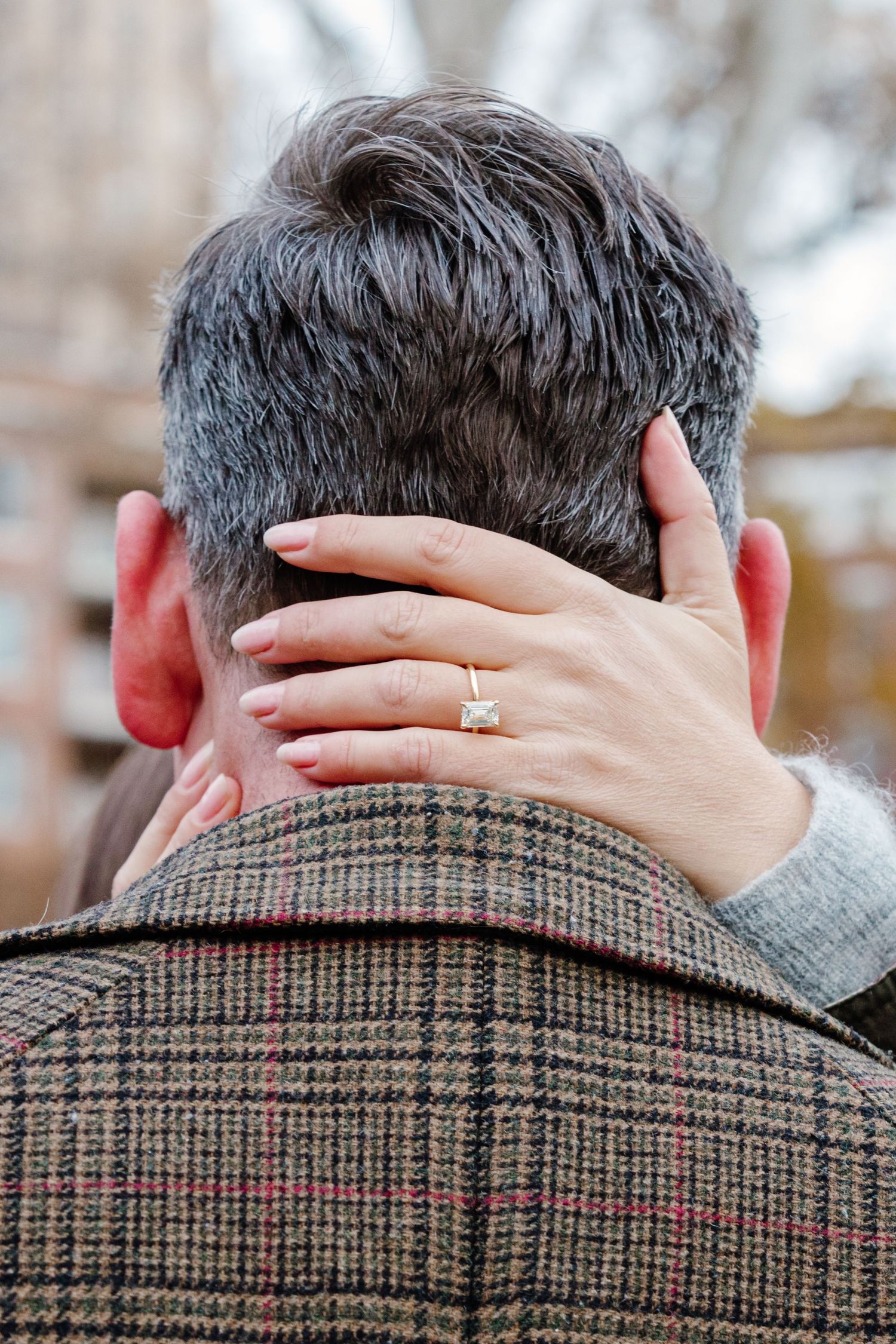 Close-up of woman's hand with gold engagement ring resting on man's neck, wearing plaid coat outdoors.