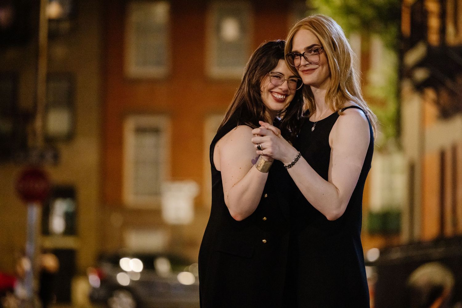 Two women in black dresses embracing and smiling on a warmly lit NYC street at night.