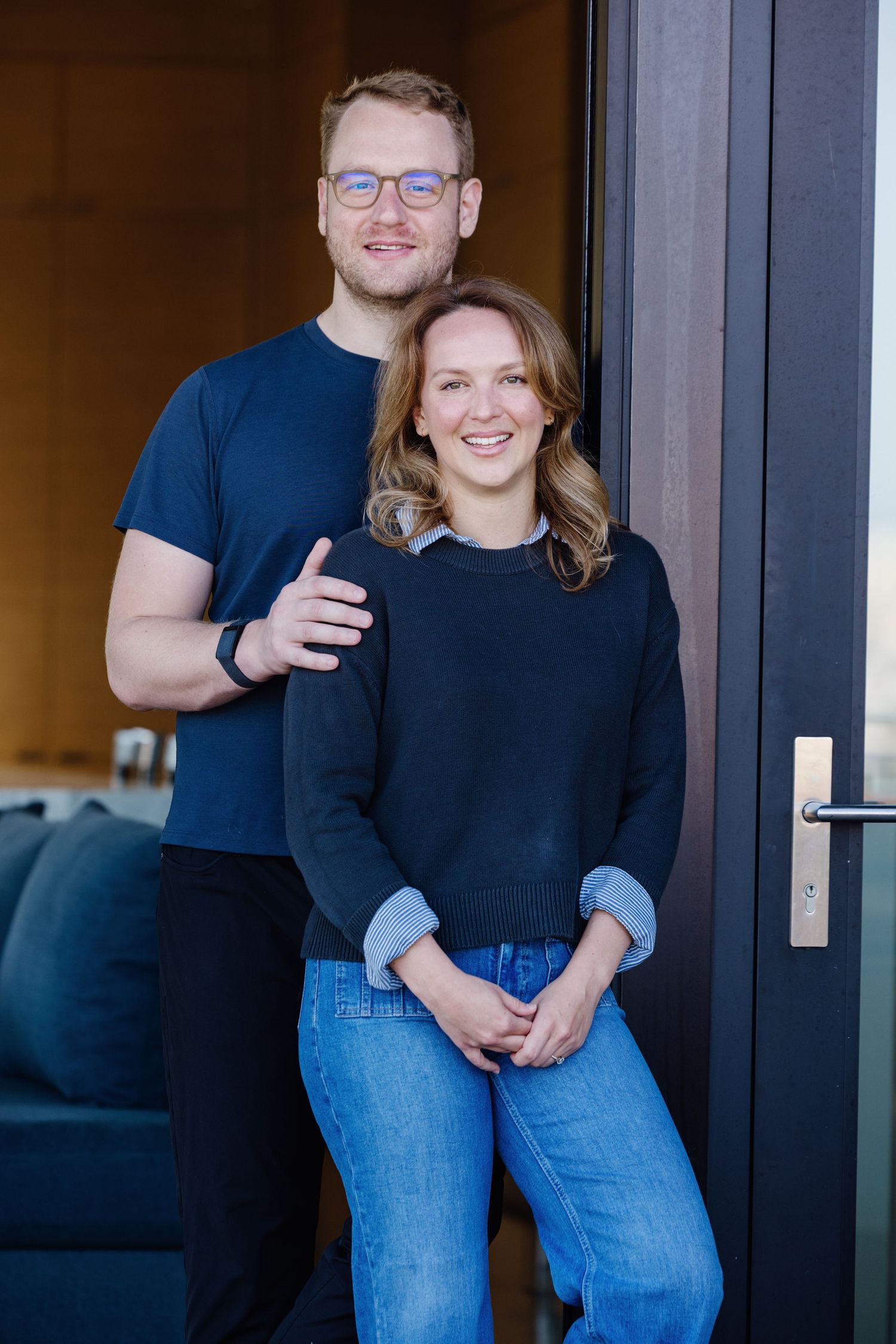Smiling couple posing together in a modern home doorway, man wearing glasses and woman in blue jeans.