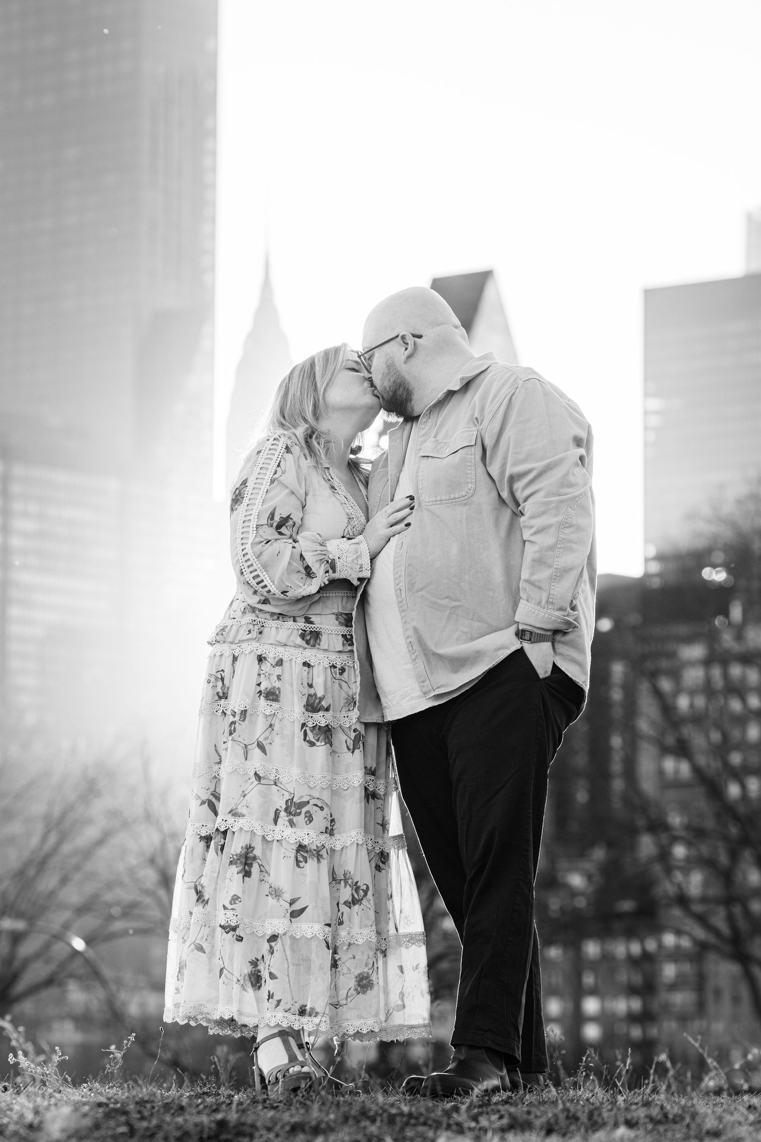 Black and white photo of a couple kissing in front of a city skyline with tall buildings in the background.