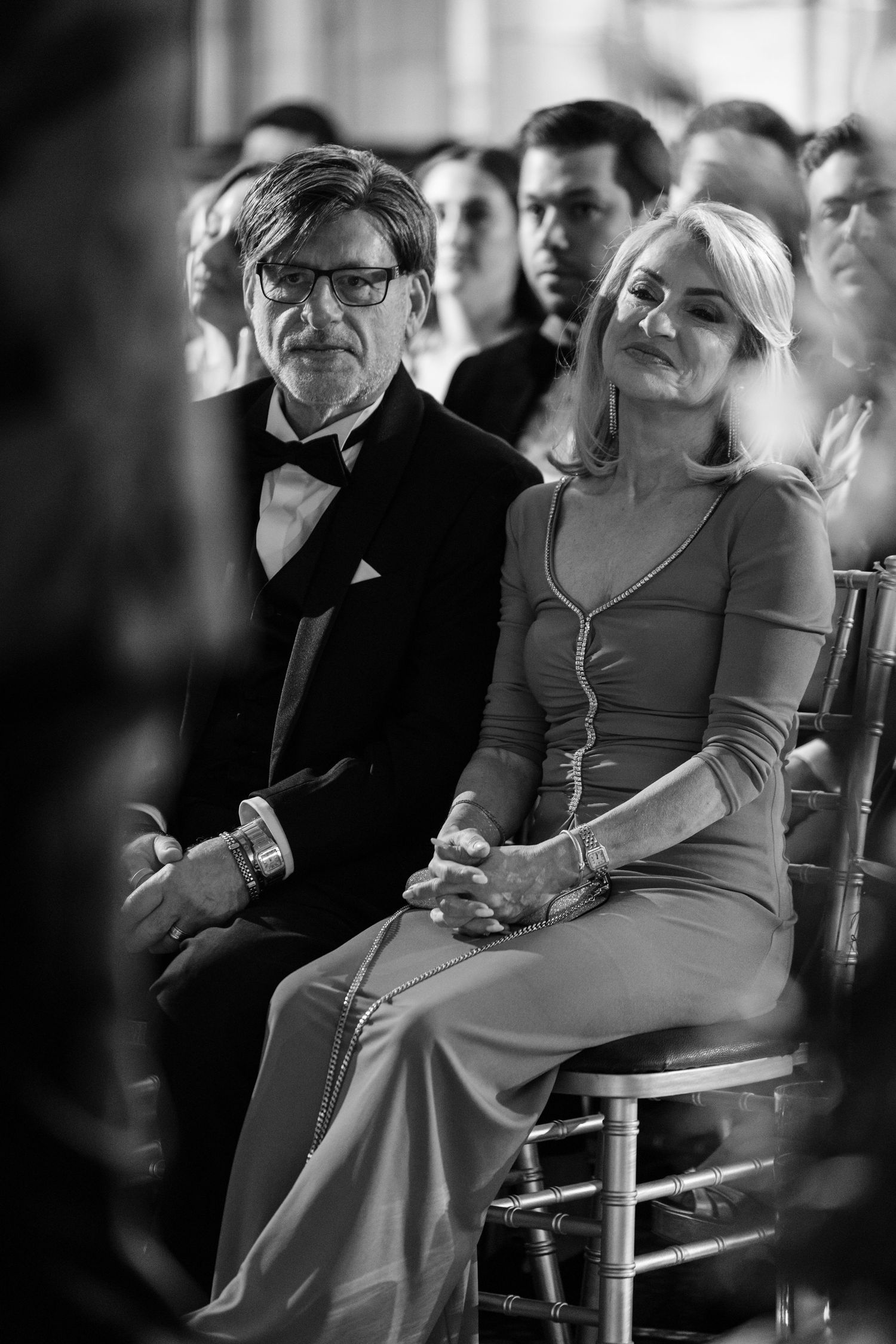Elegant couple in formal attire sits attentively as wedding ceremony guests in black and white photo.