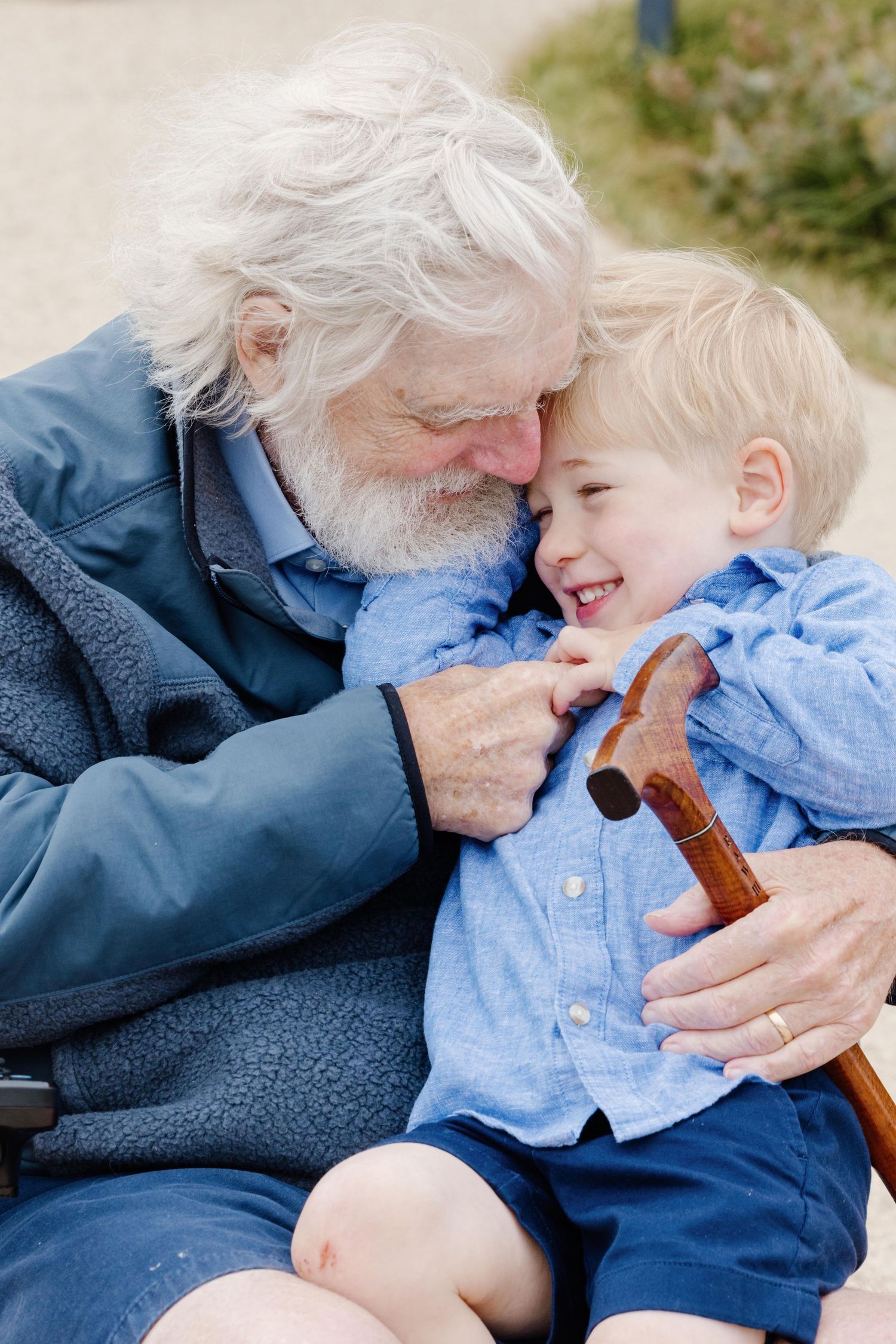 Elderly grandfather with white beard hugs laughing young blonde boy holding a wooden cane outdoors.