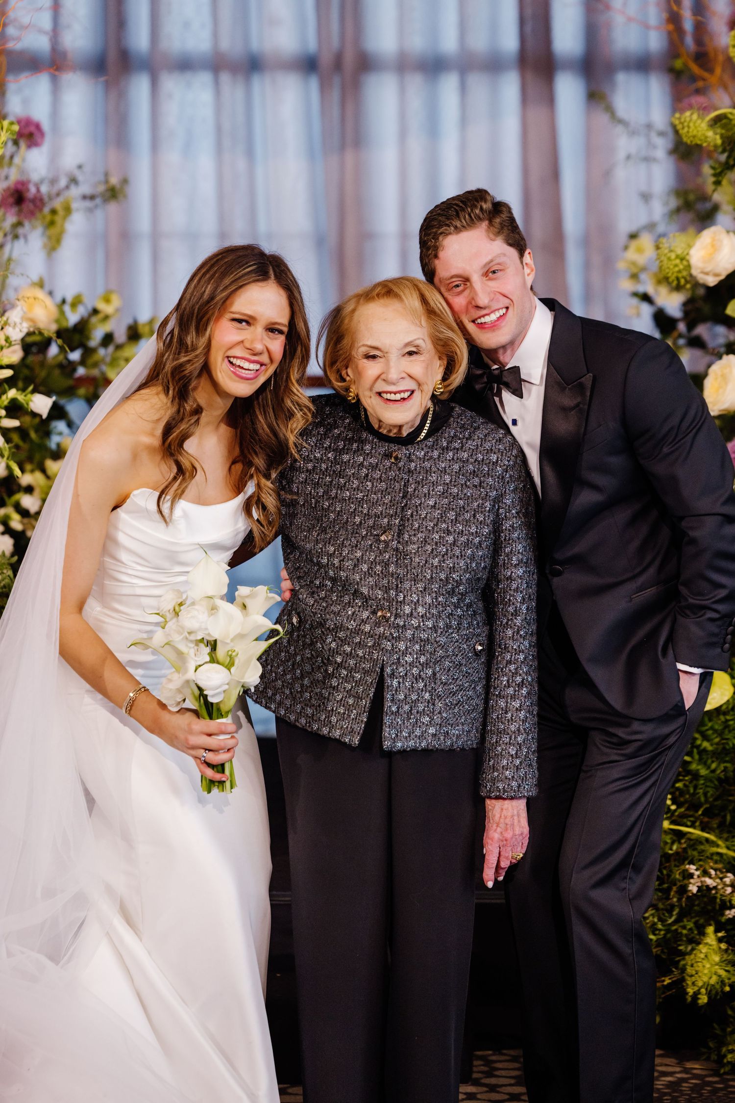 Bride in white gown and groom in tuxedo posing with elderly woman at wedding reception.