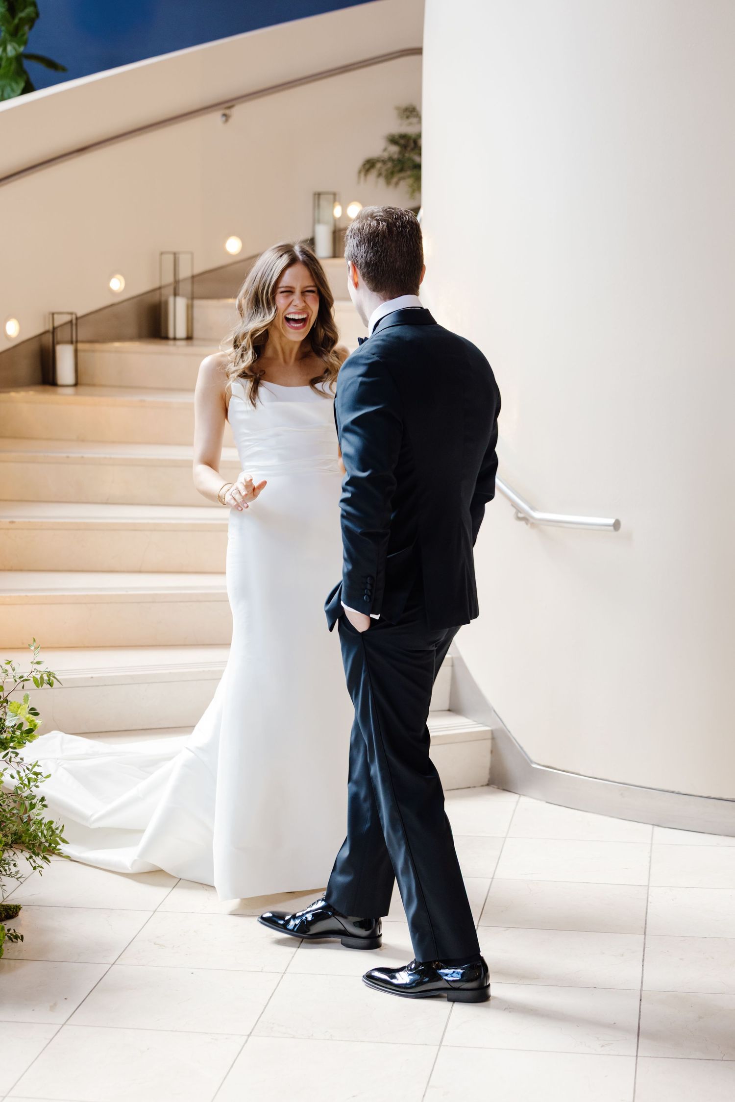 Bride in white mermaid gown laughs joyfully during first look moment with groom in black tuxedo.