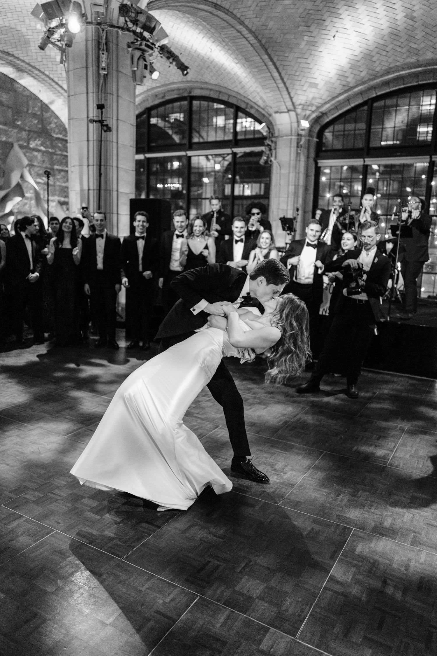 Groom dramatically dips bride during first dance as guests cheer in a grand arched reception hall.