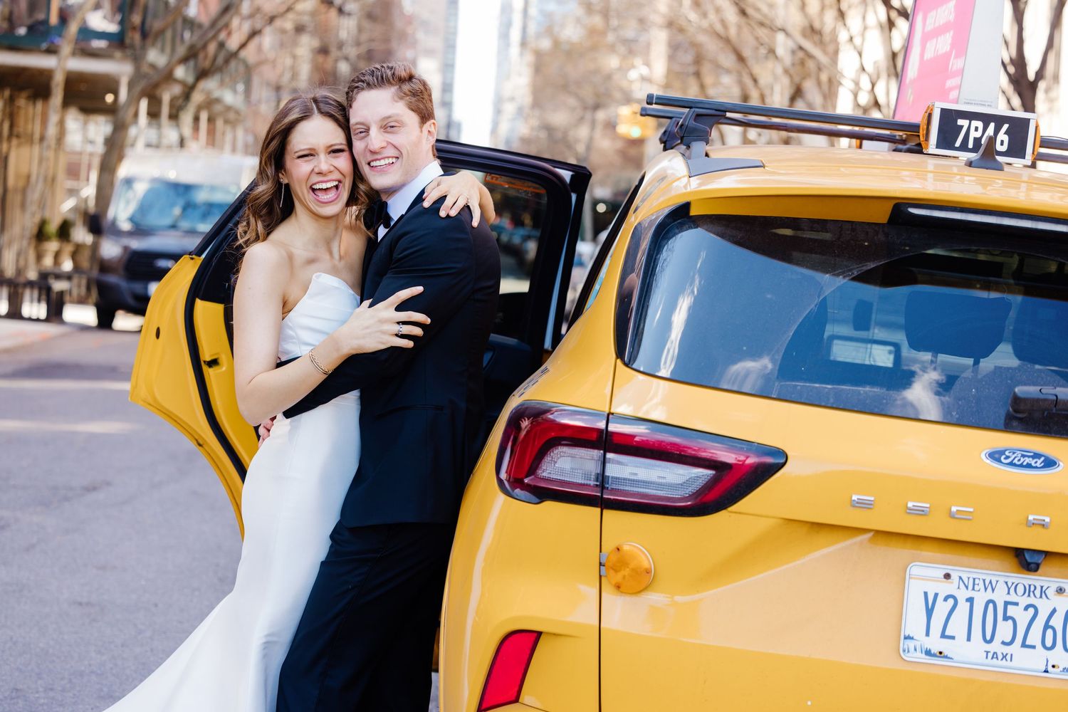 Laughing bride and groom in wedding attire pose beside a yellow New York City taxi cab on city street.