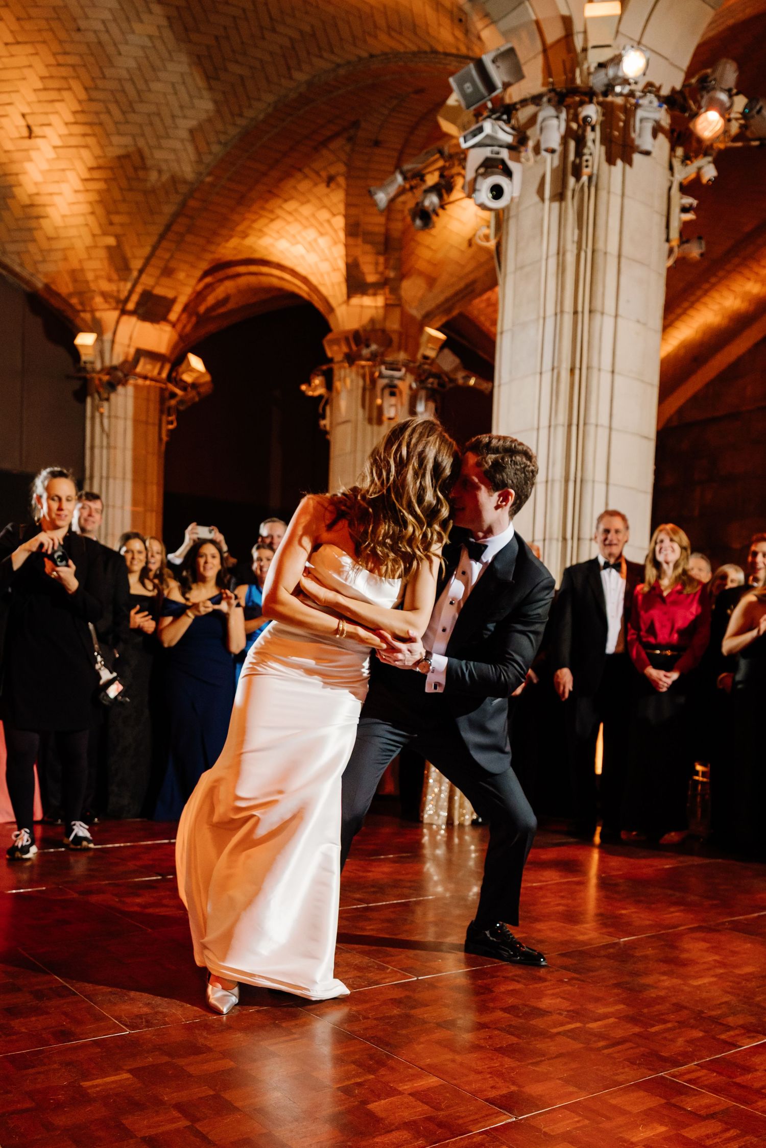 Couple shares a joyful first dance in a stunning brick-arched ballroom with glowing warm lighting.