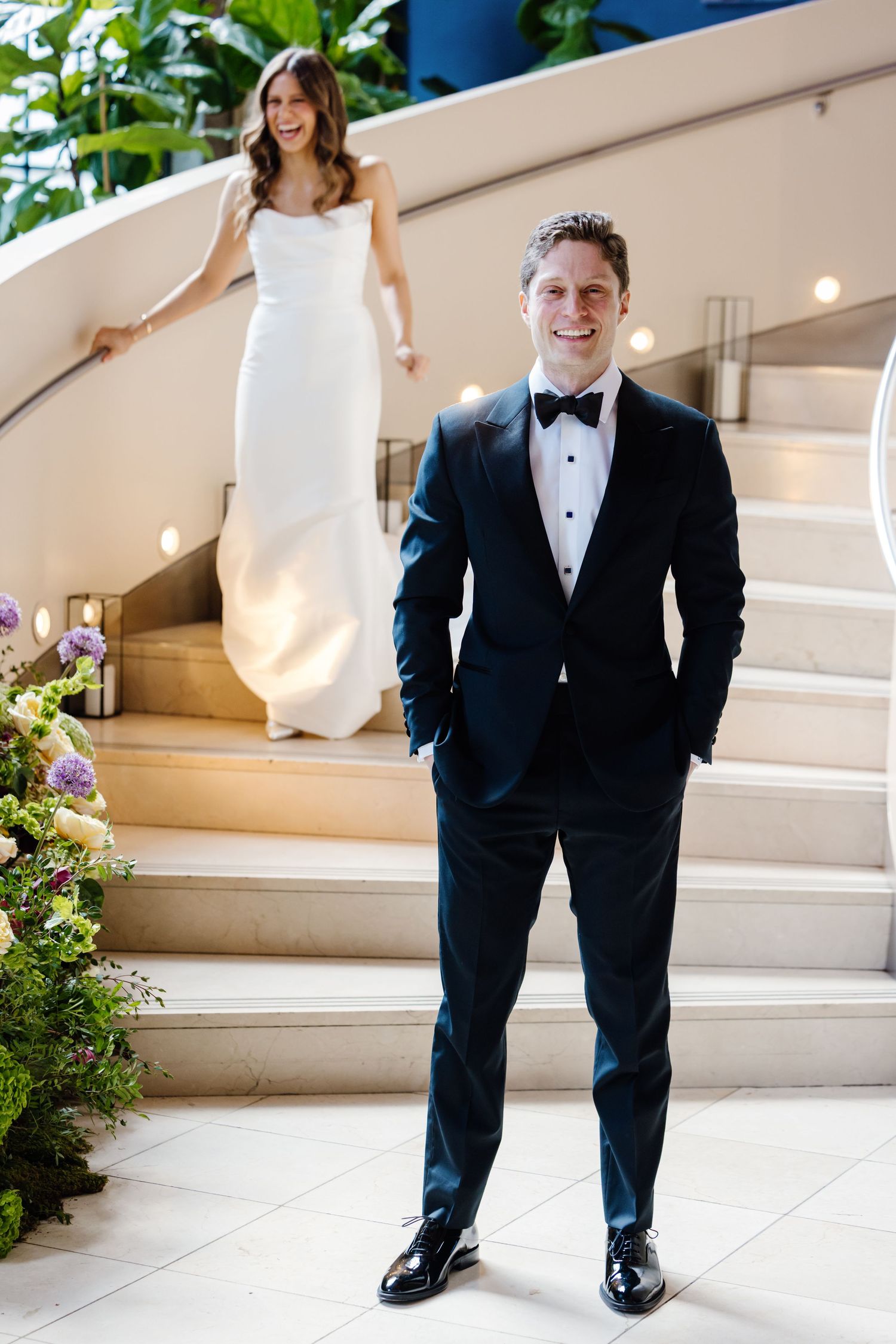 Groom in black tuxedo smiles at camera as bride descends elegant curved staircase in sleek white gown.