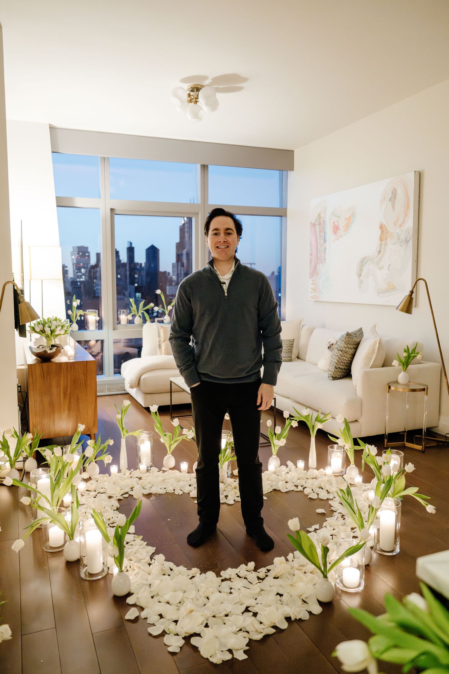Man standing inside a heart-shaped arrangement of white rose petals, tulips, and candles in a modern apartment.