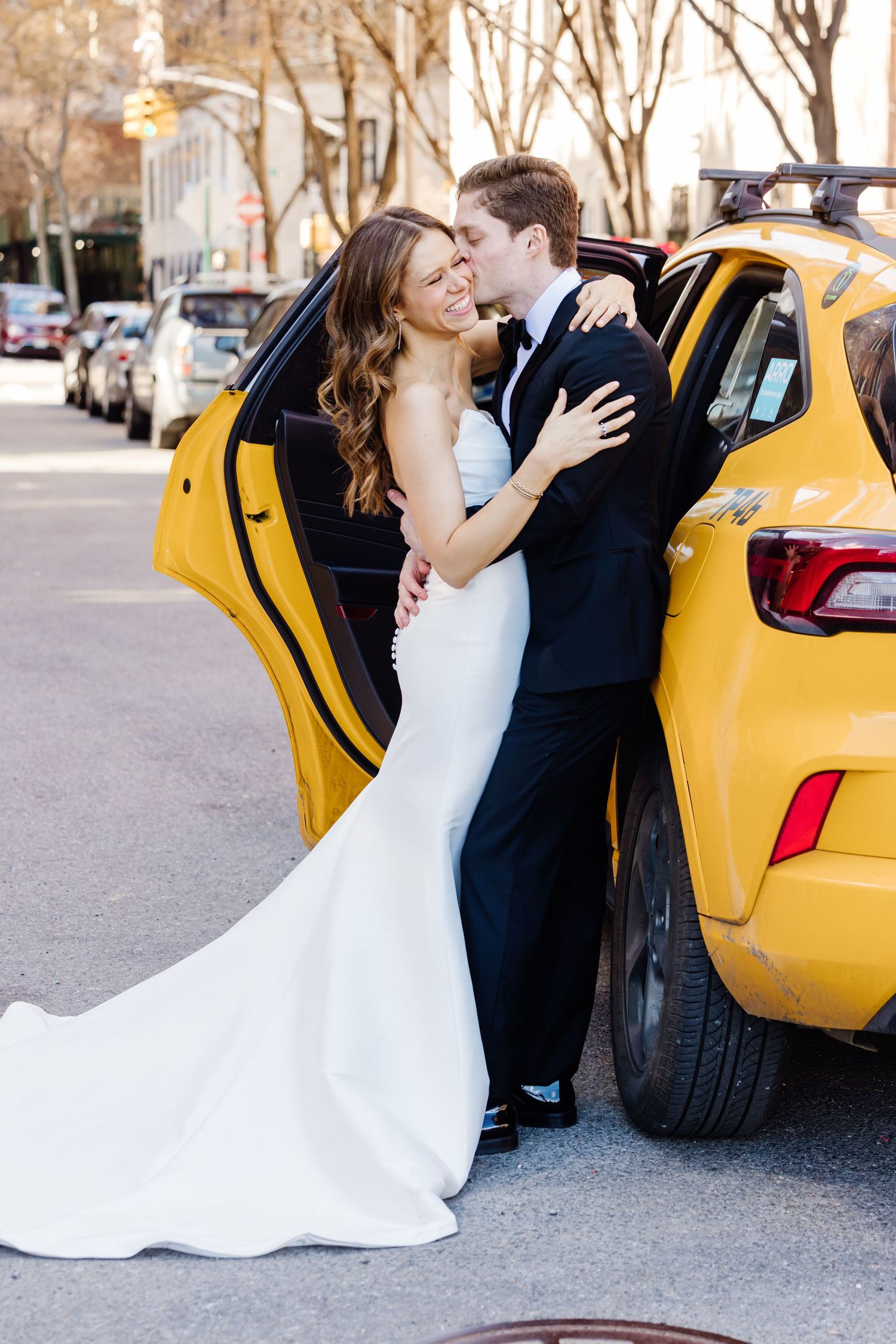 Bride and groom laughing together leaning against a yellow NYC taxi cab on a tree-lined street.