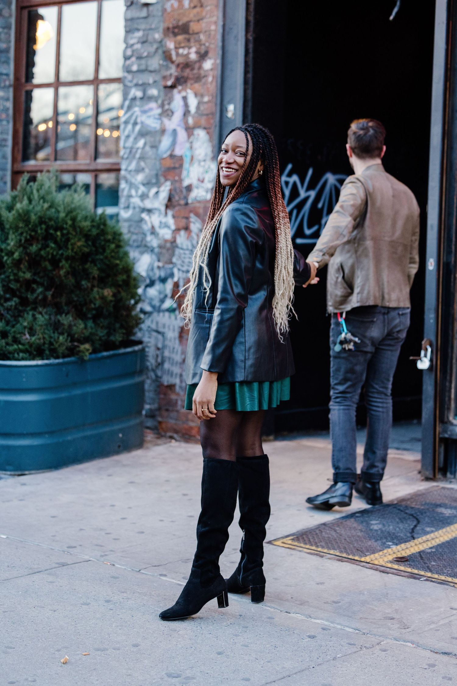 Smiling woman with long braids in leather jacket and green skirt posing on NYC street.