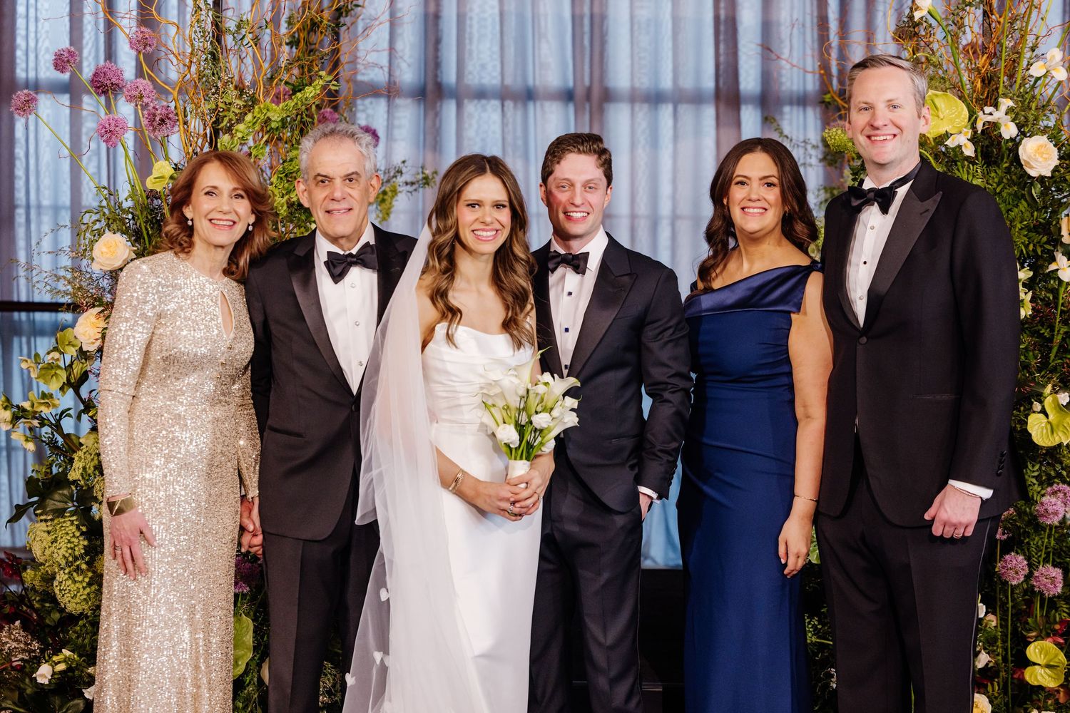 Six formally dressed wedding guests and newlyweds smiling together before colorful floral backdrop.