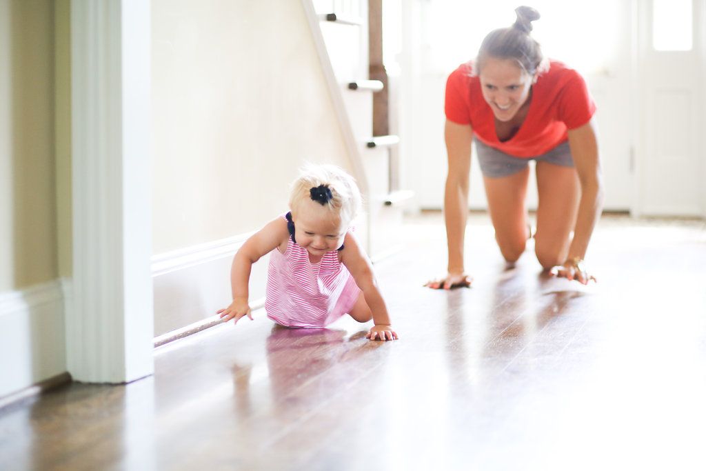Mother and baby girl playing on hardwood floor together, with mom doing push-ups as toddler crawls nearby.