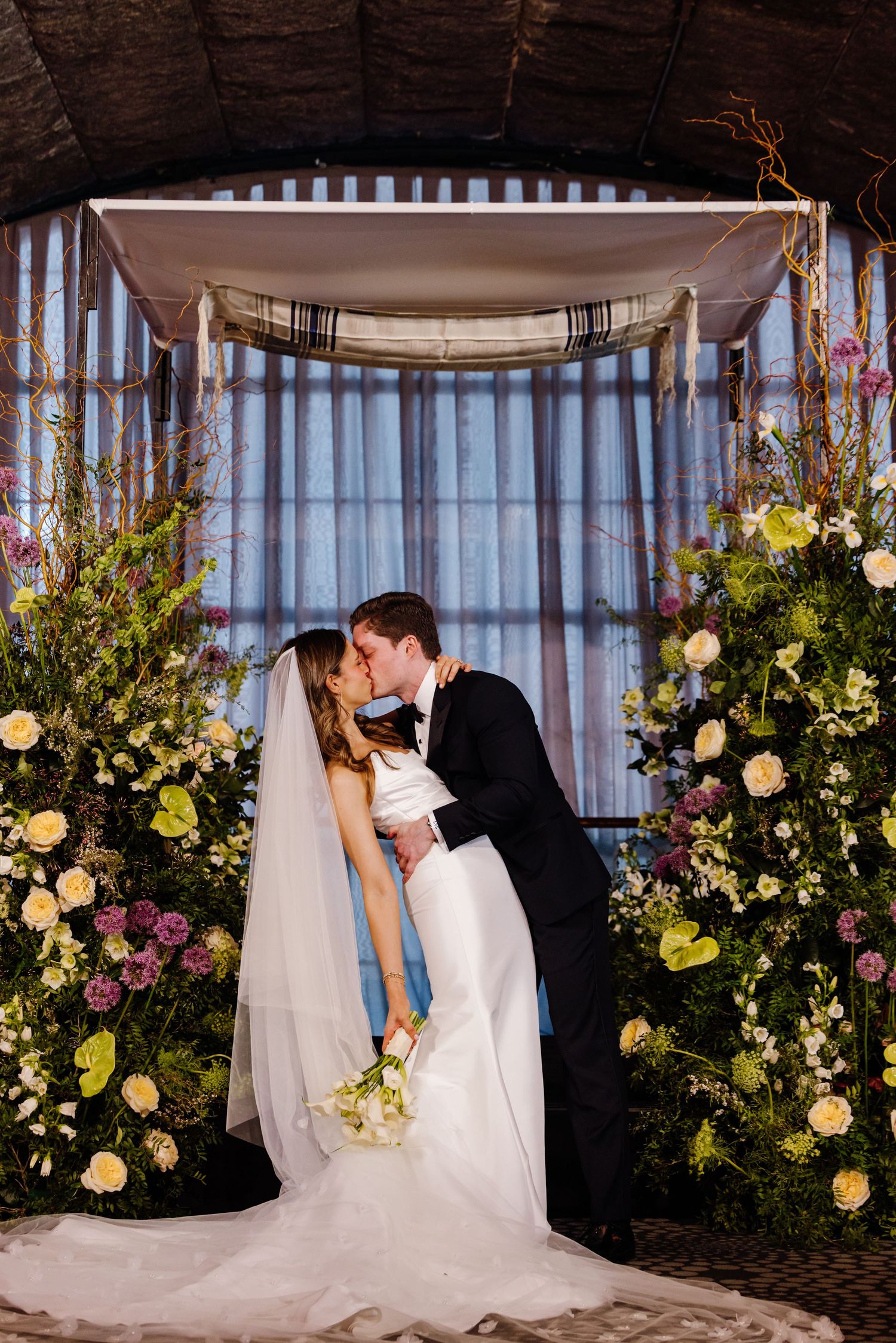 Bride and groom sharing their first kiss under a lush floral chuppah during their wedding ceremony.