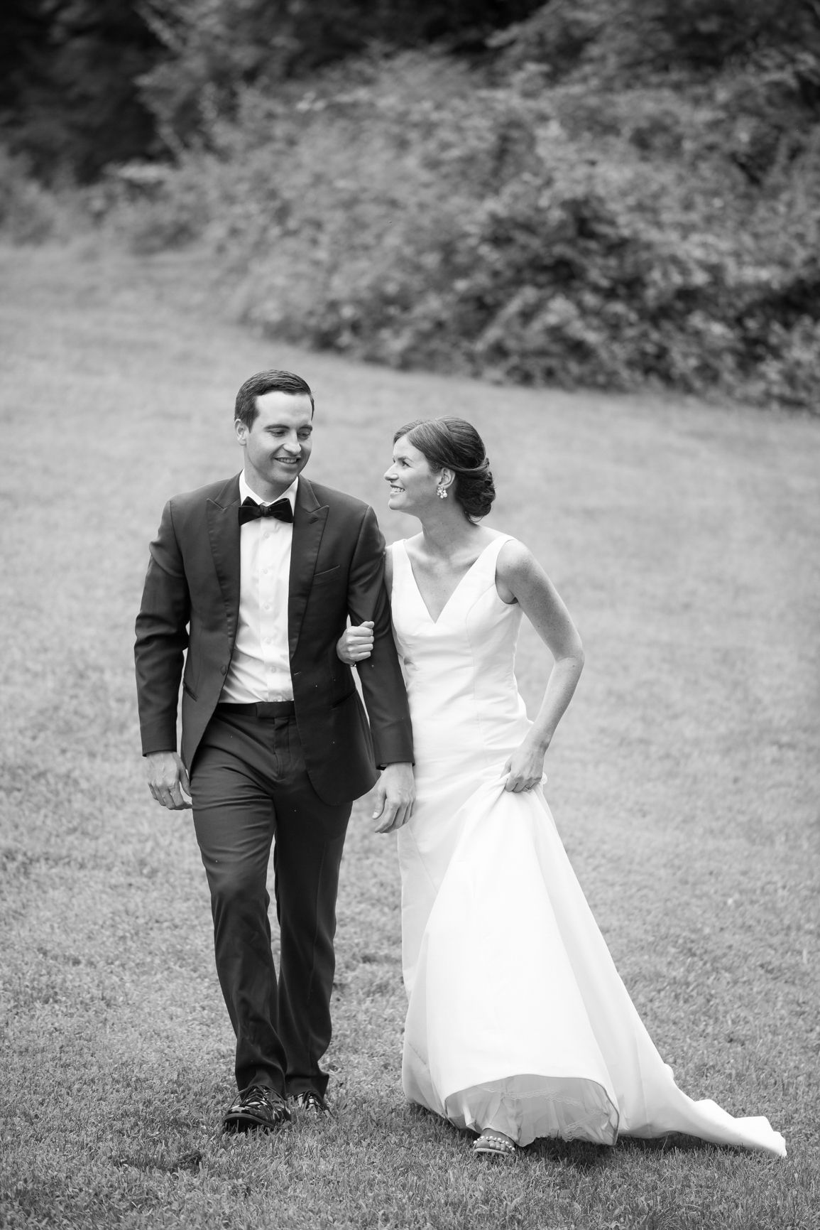 Black and white photo of bride and groom walking arm in arm on a grassy lawn.