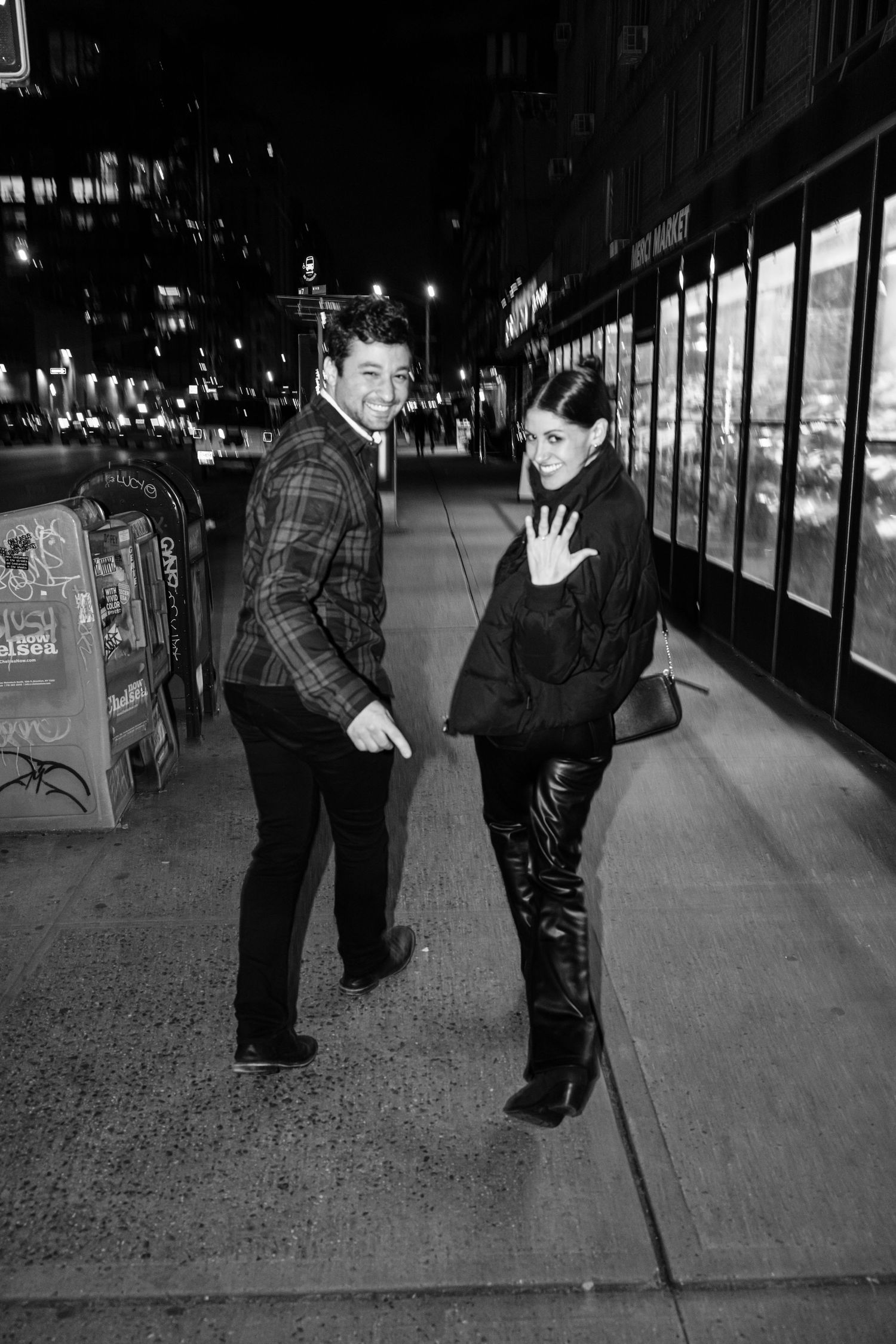 Newly engaged couple posing on NYC sidewalk at night, woman showing off engagement ring.