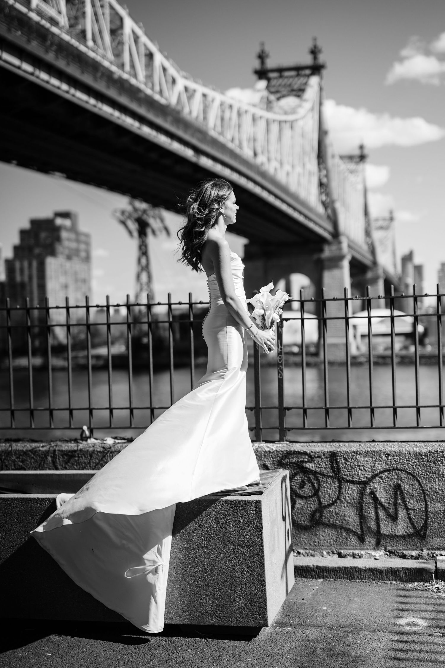 Black and white photo of a bride in a fitted gown holding bouquet beside the Queensboro Bridge.