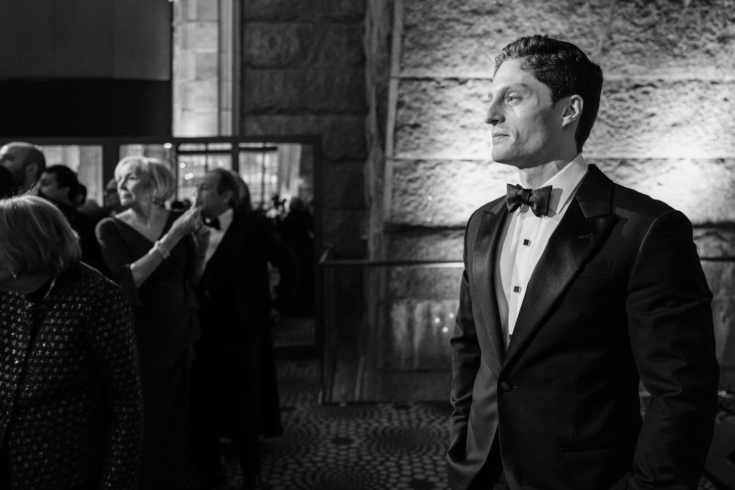 Dapper groom in black tuxedo and bow tie stands outside elegant wedding venue in black and white photo.