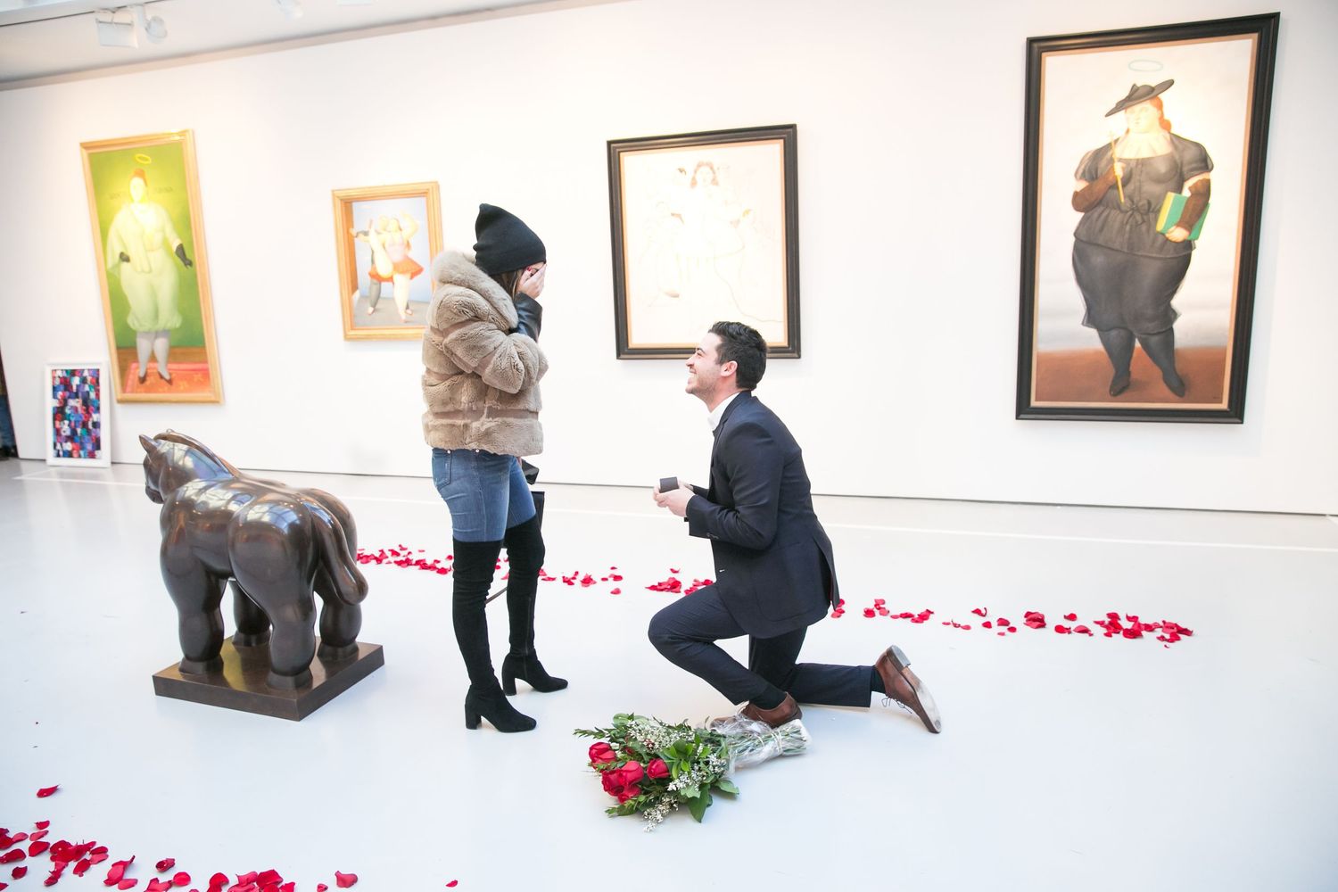 Man proposing in art gallery with rose petals on floor and bouquet of roses.
