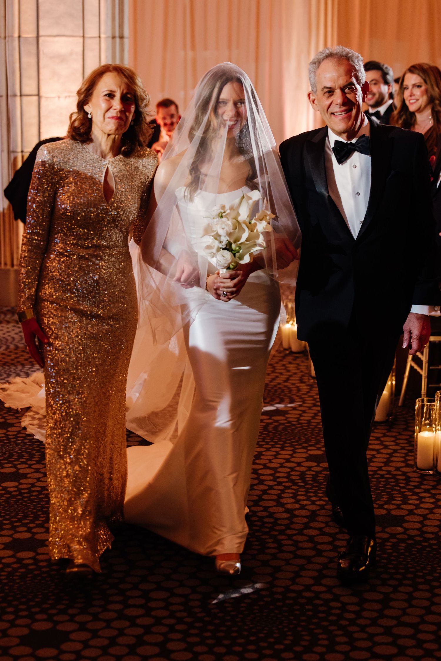 Bride in white gown and veil walks down aisle escorted by parents in formal attire at wedding.