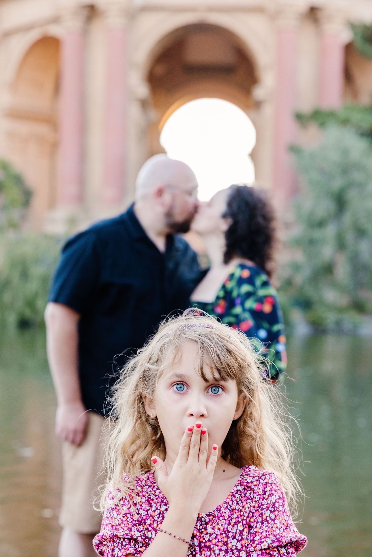 Young girl blows a kiss while parents kiss behind her at the Palace of Fine Arts rotunda.