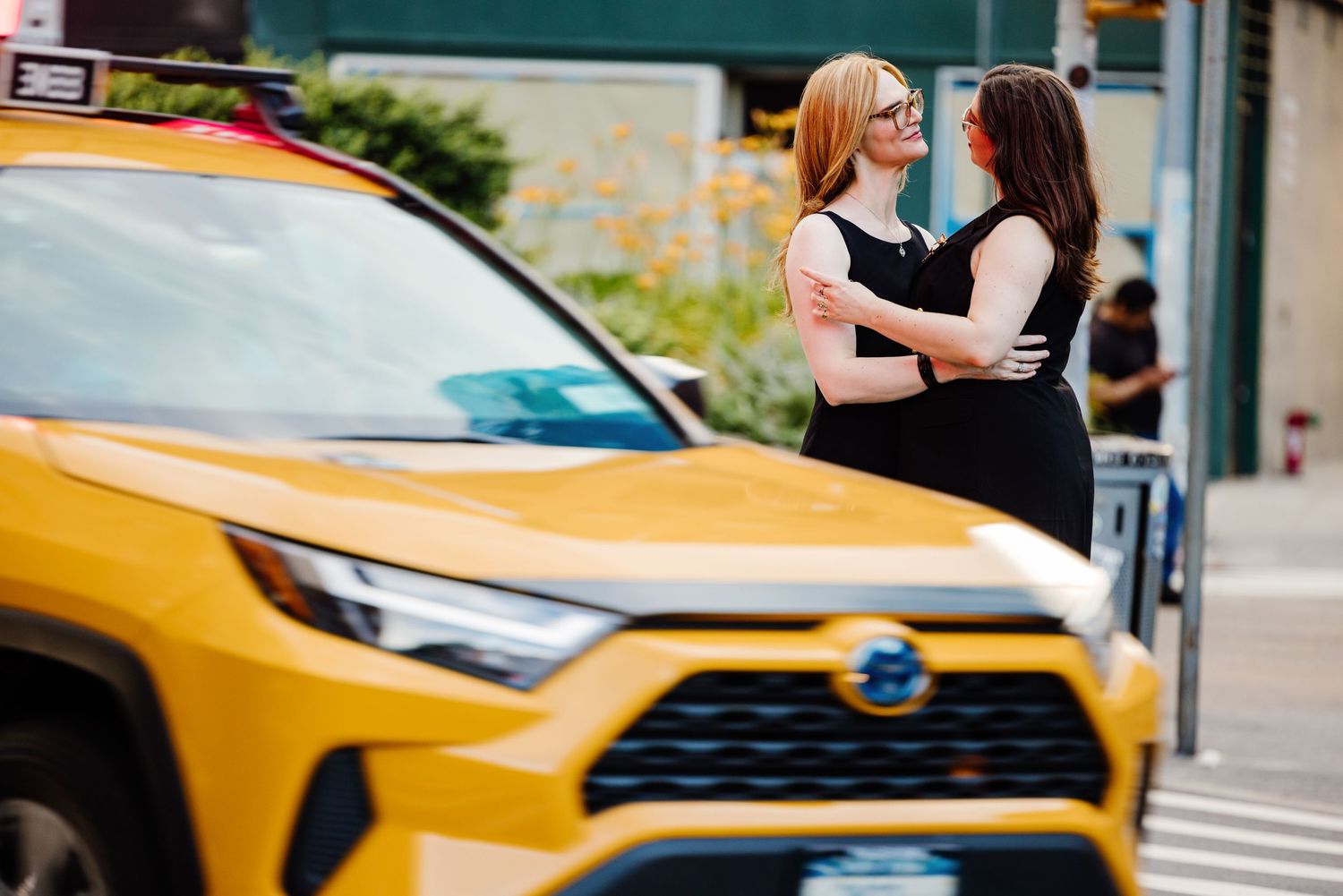 Two women embracing near a yellow NYC taxi cab on a city street.