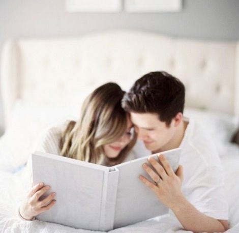 Couple lying in bed together reading a white hardcover photo album.