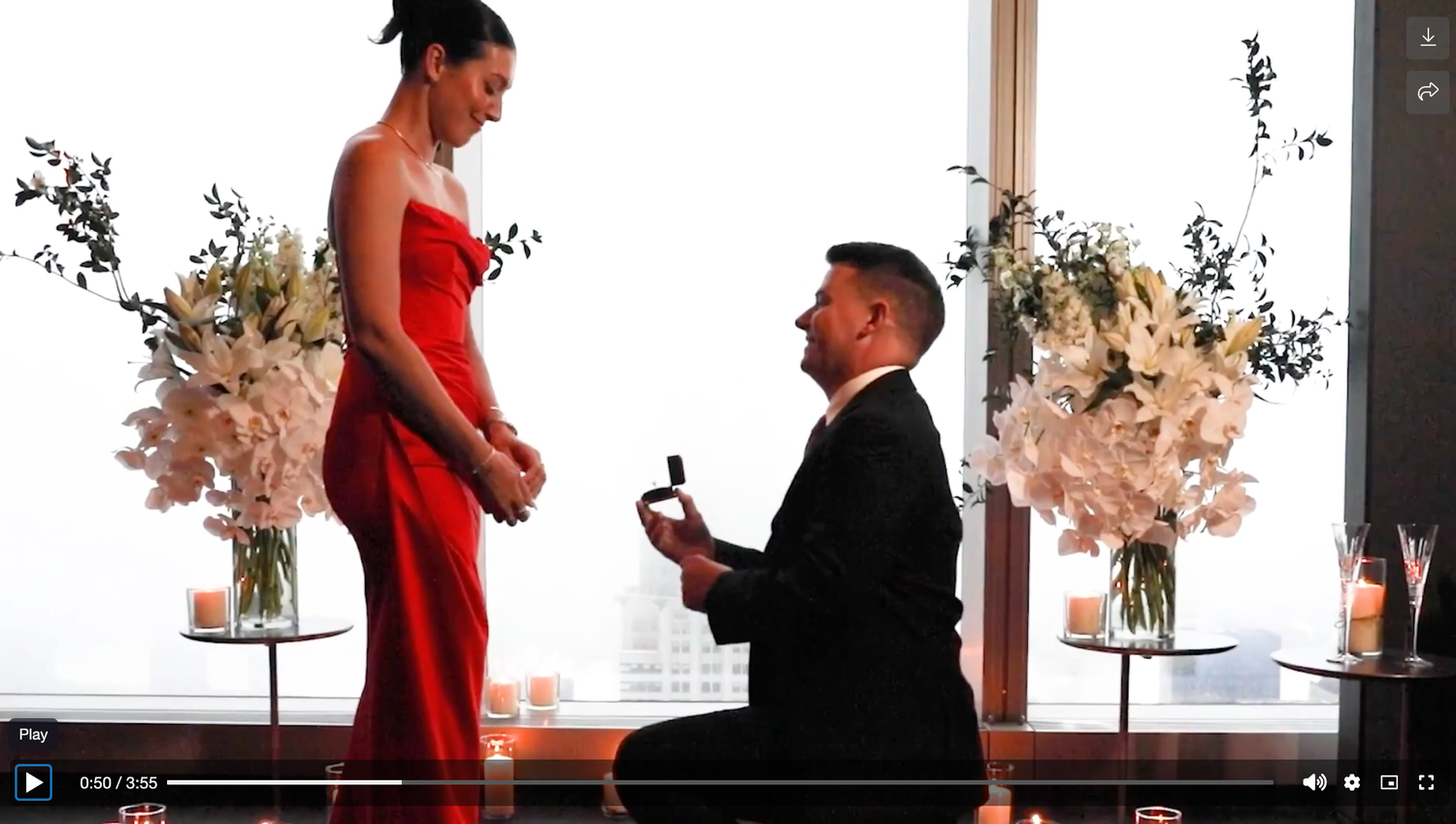 Man proposing to woman in red dress with ring box, romantic setting with white flowers.