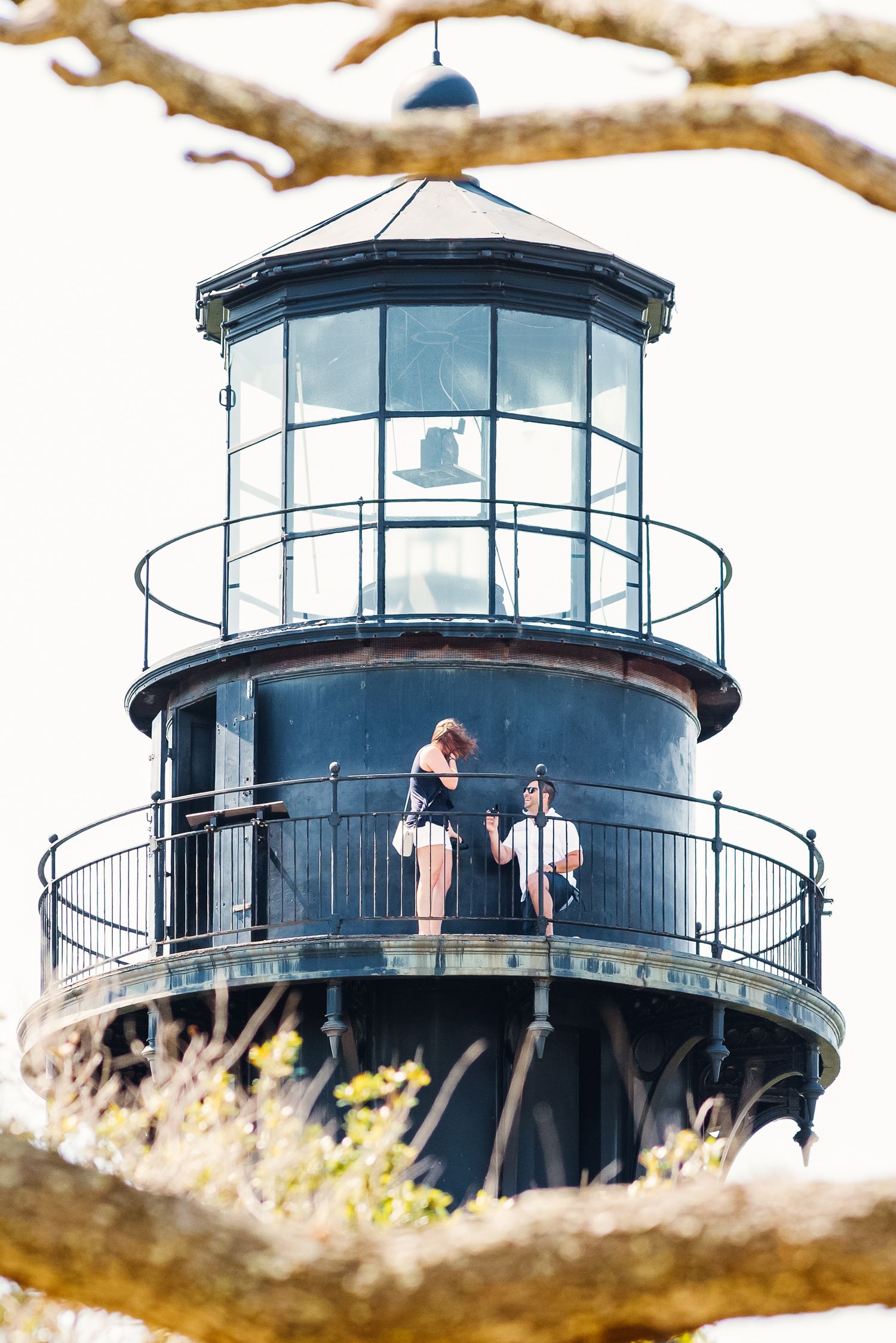 Couple standing together on the circular balcony of a tall dark lighthouse framed by bare tree branches.