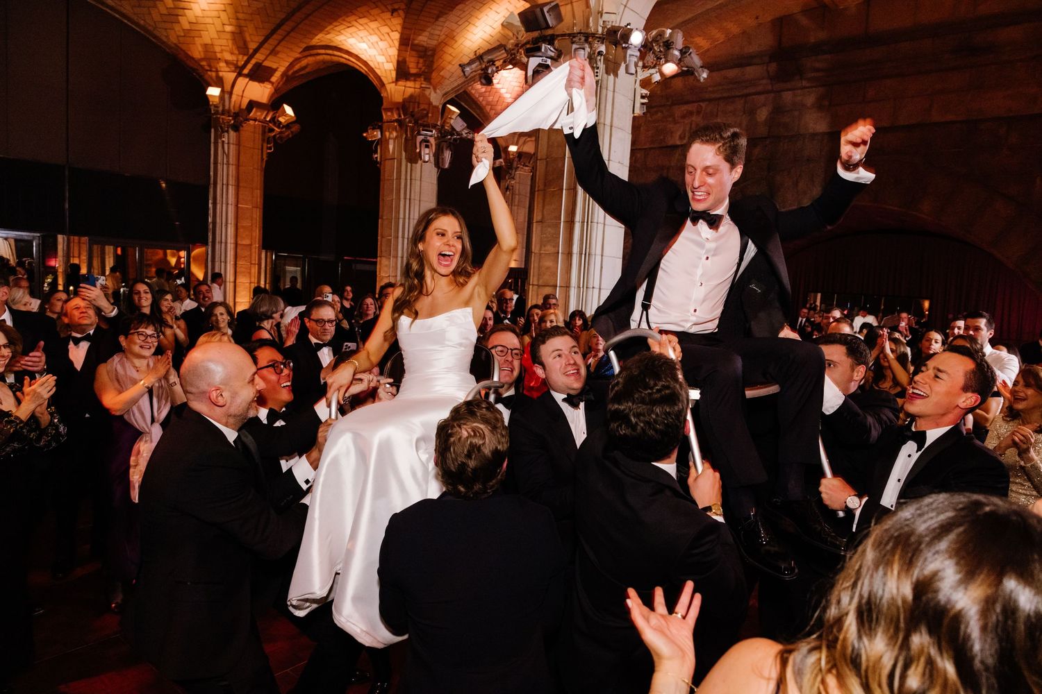 Bride and groom lifted by guests during joyful wedding reception hora dance in elegant ballroom.