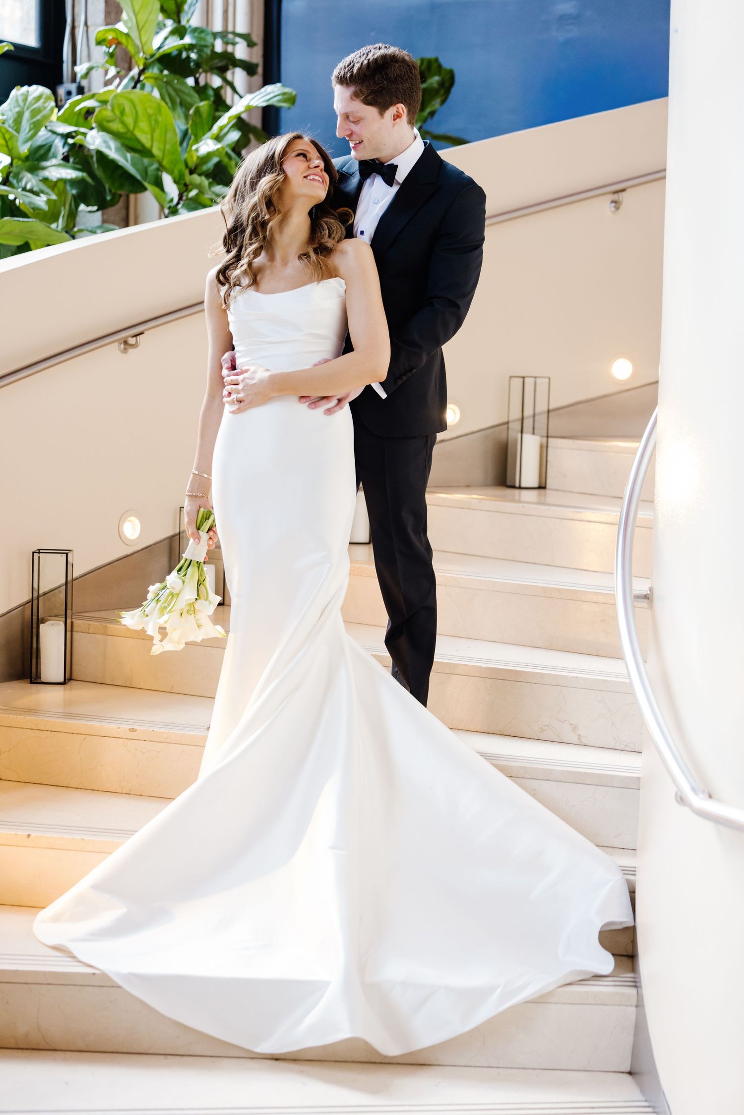 Groom embraces bride in strapless white mermaid gown on staircase as she holds white floral bouquet.