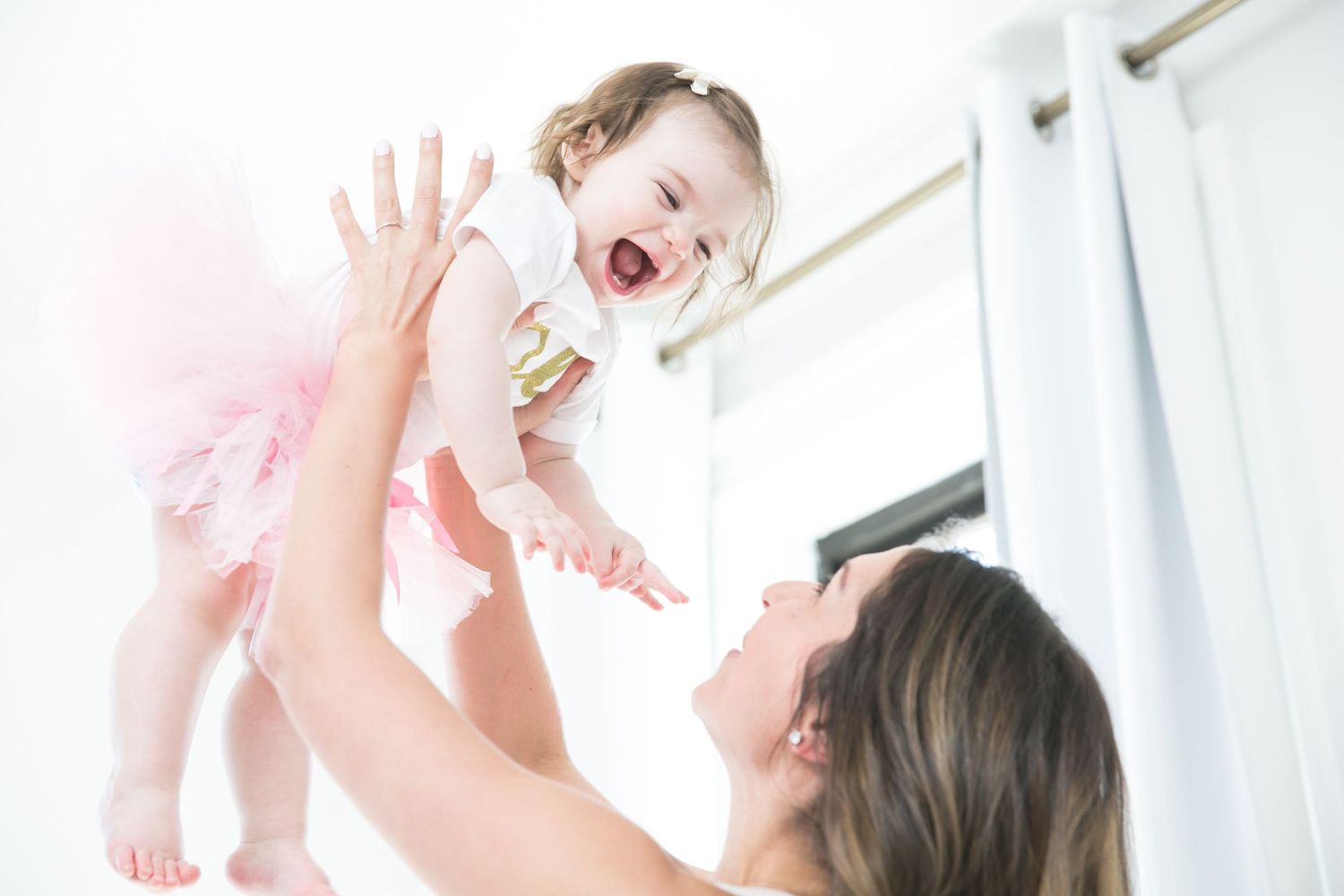A joyful mother lifts her laughing baby girl in a pink tutu dress in a bright white room.