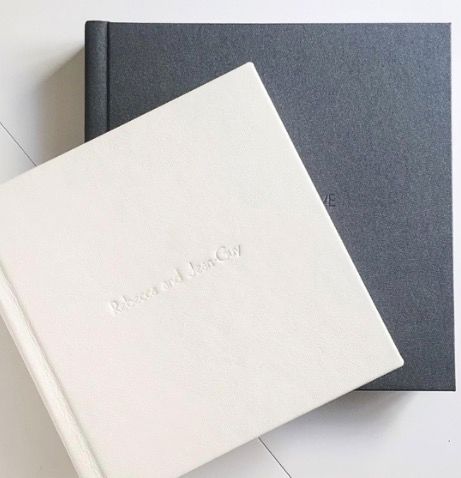 Two elegant hardcover photo albums in white linen and dark gray fabric with embossed text on a white surface.
