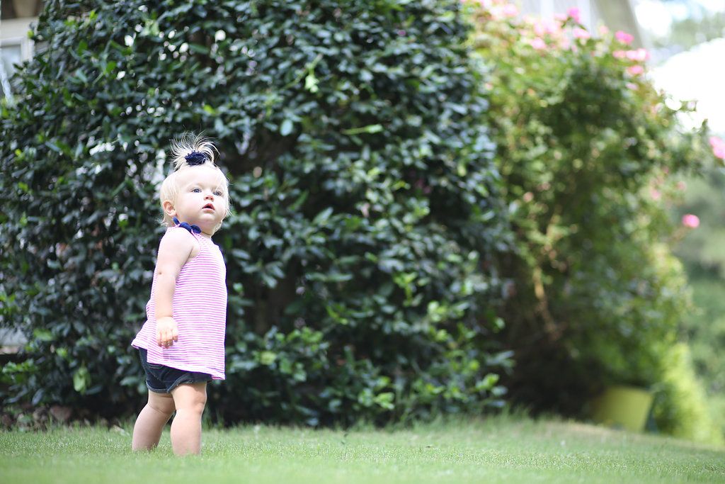 Cute toddler girl in pink dress with sunglasses playing on green grass in a garden.