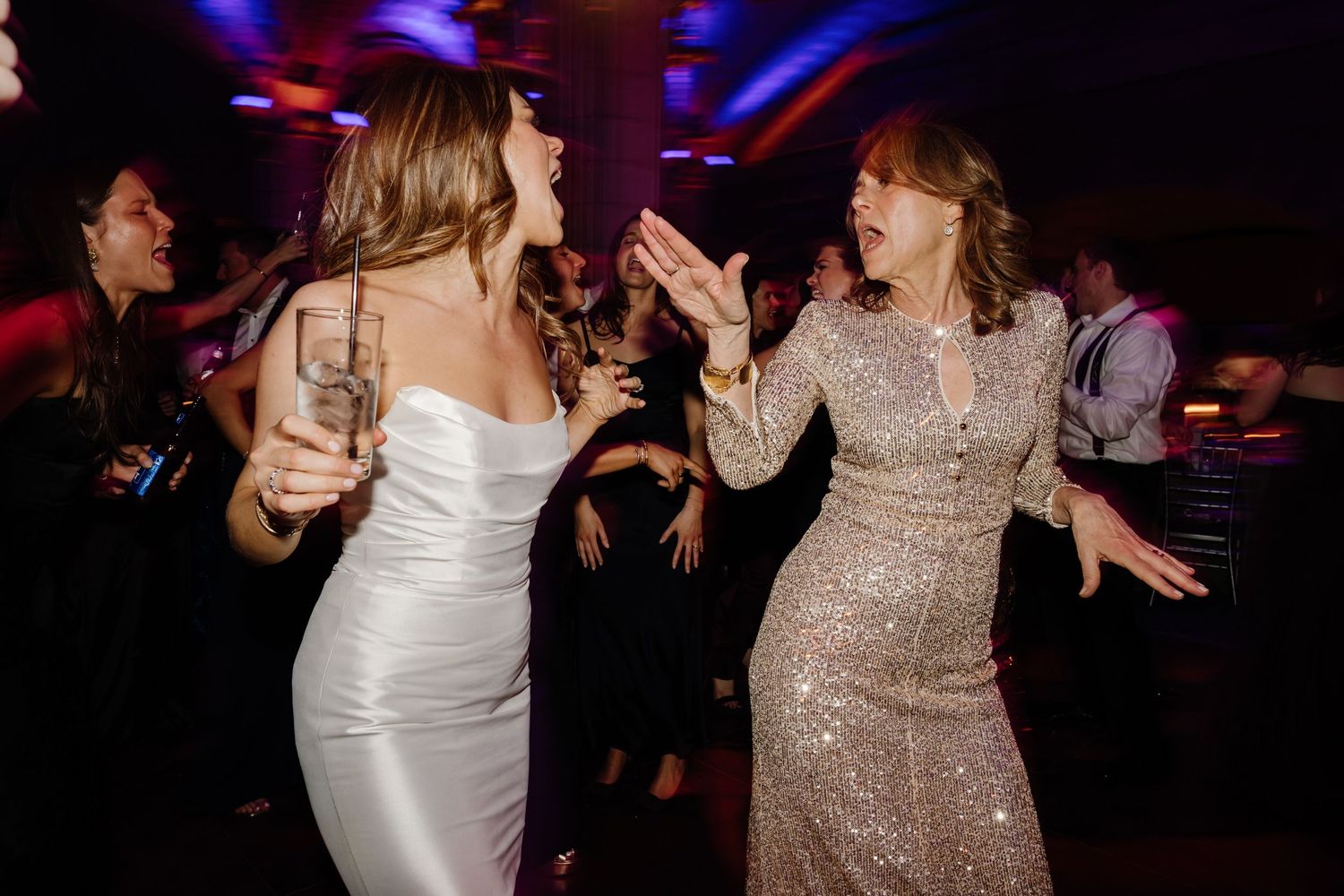 Bride in white gown and guest in sequin dress dancing joyfully at a vibrant wedding reception.