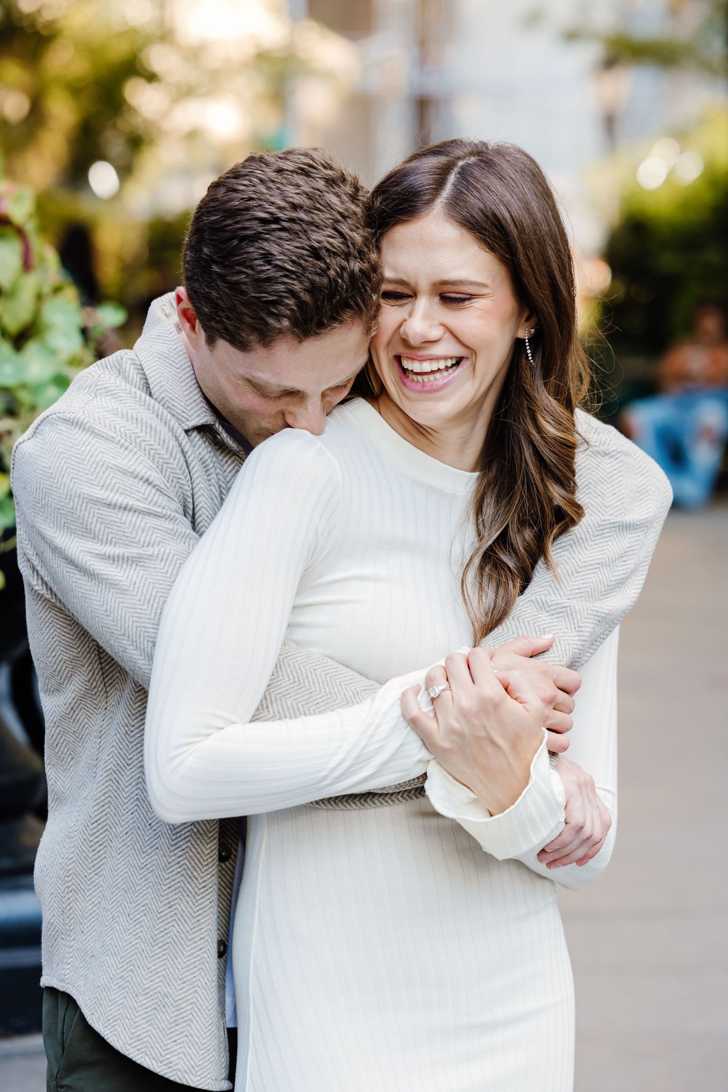 Happy couple embracing outdoors, woman in white dress laughing during engagement photo session.