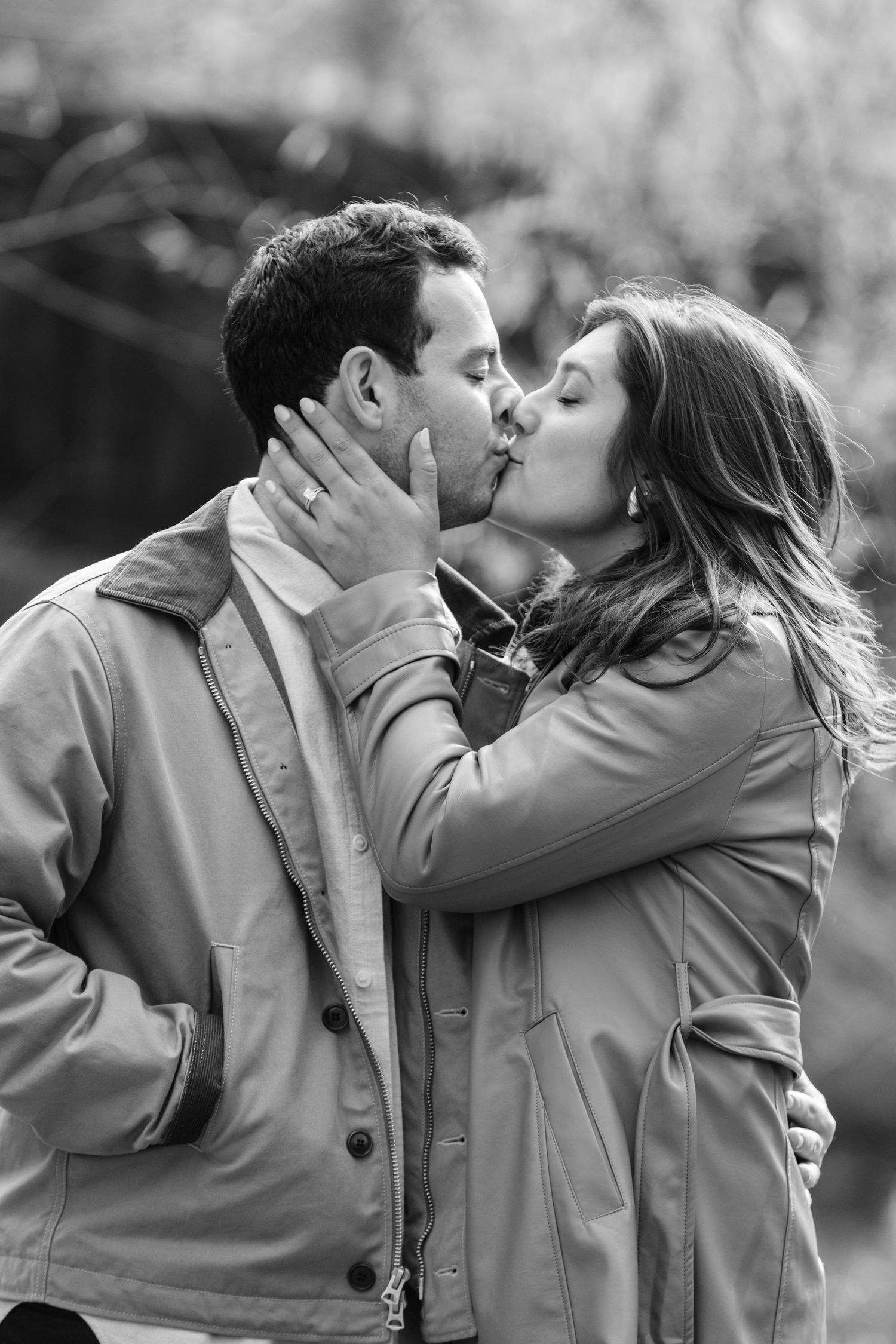 Black and white photo of couple sharing romantic kiss outdoors wearing matching trench coats.
