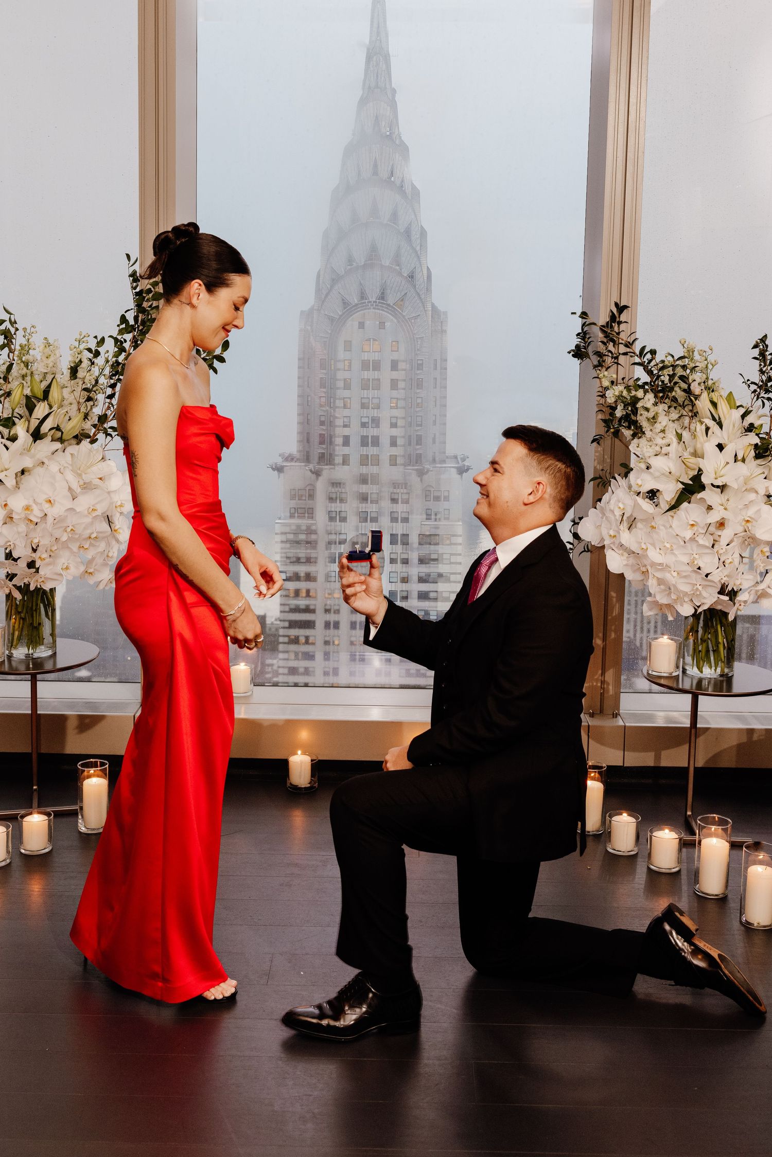 Man proposing on one knee to woman in red gown with Chrysler Building view backdrop.