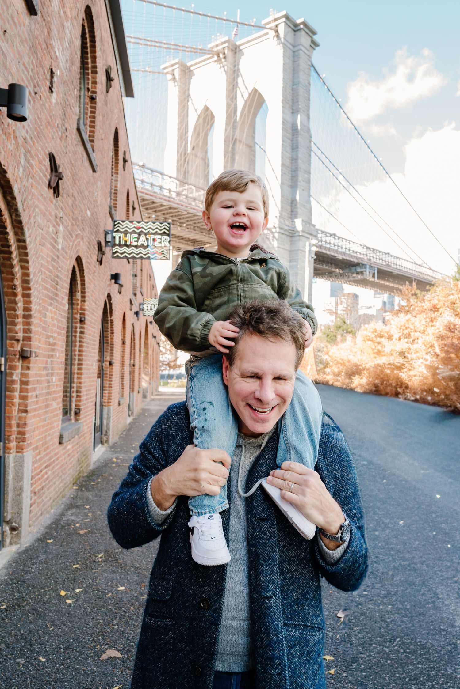 Smiling toddler sits on fathers shoulders near Brooklyn Bridge with autumn foliage.