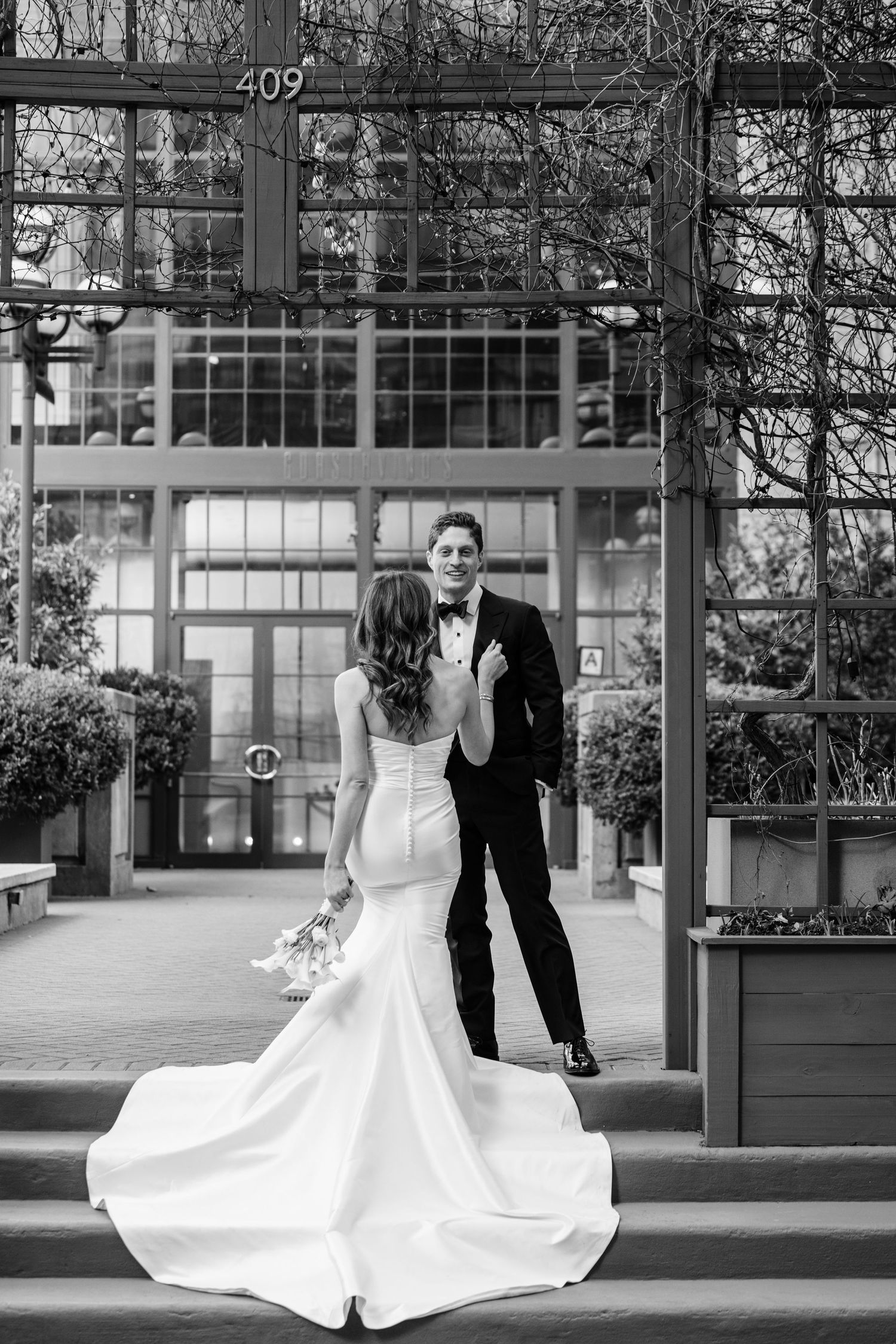 Black and white photo of a bride approaching a smiling groom on steps outside a glass building.