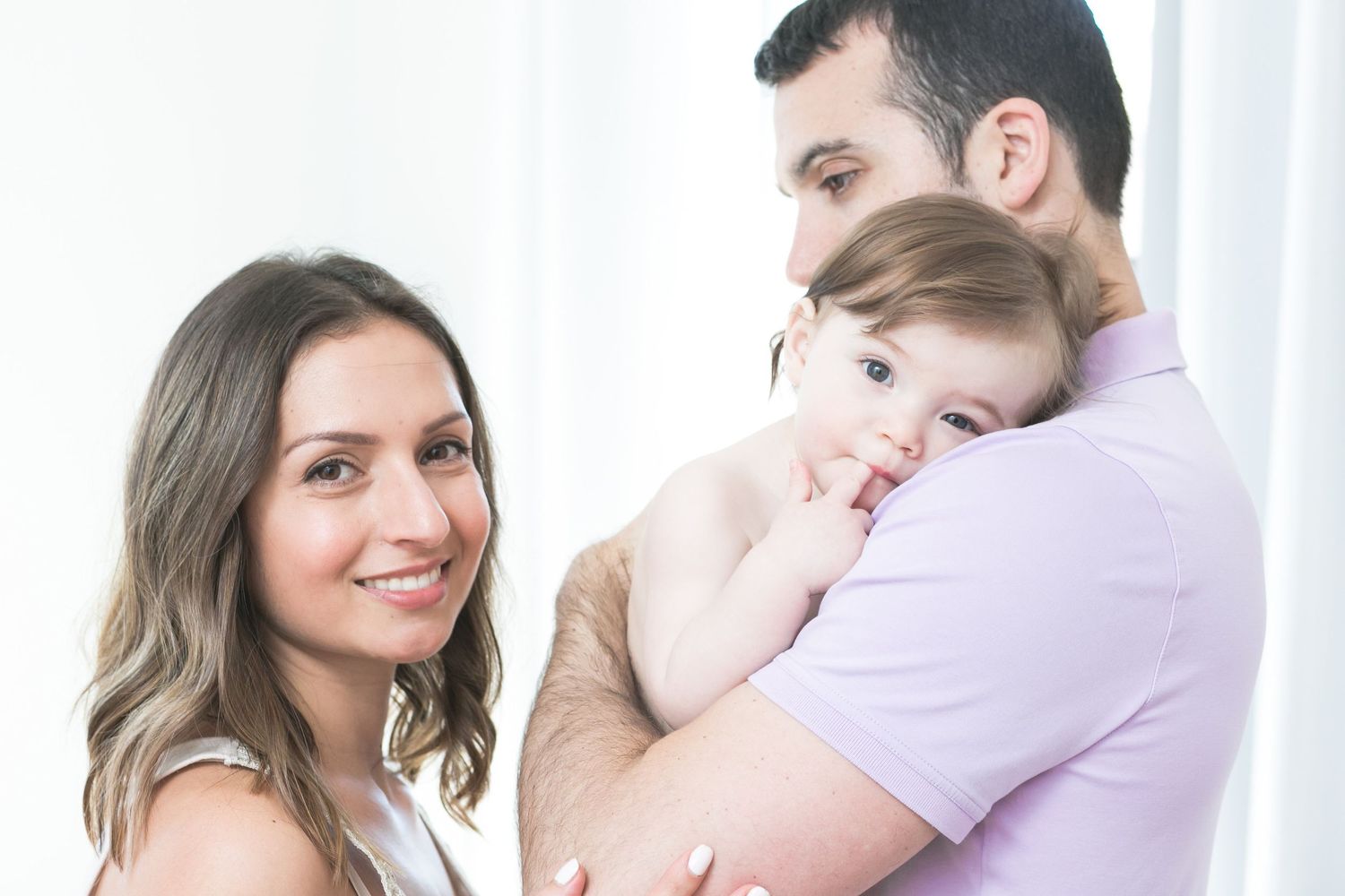 A happy young family with a mother, father, and baby posing together in a bright, white studio setting.