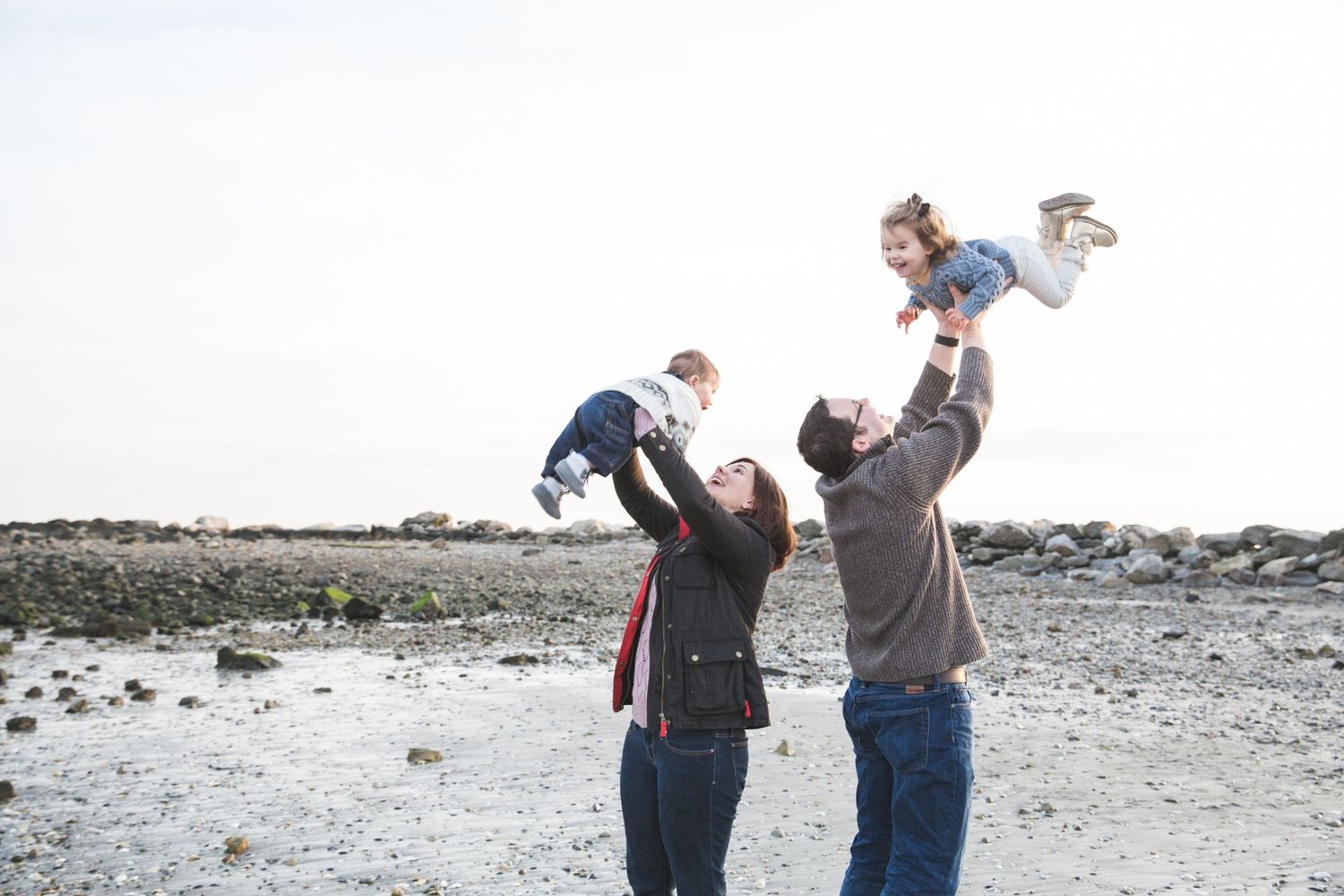 Parents joyfully lift their two young children in the air on a rocky beach under a clear sky.