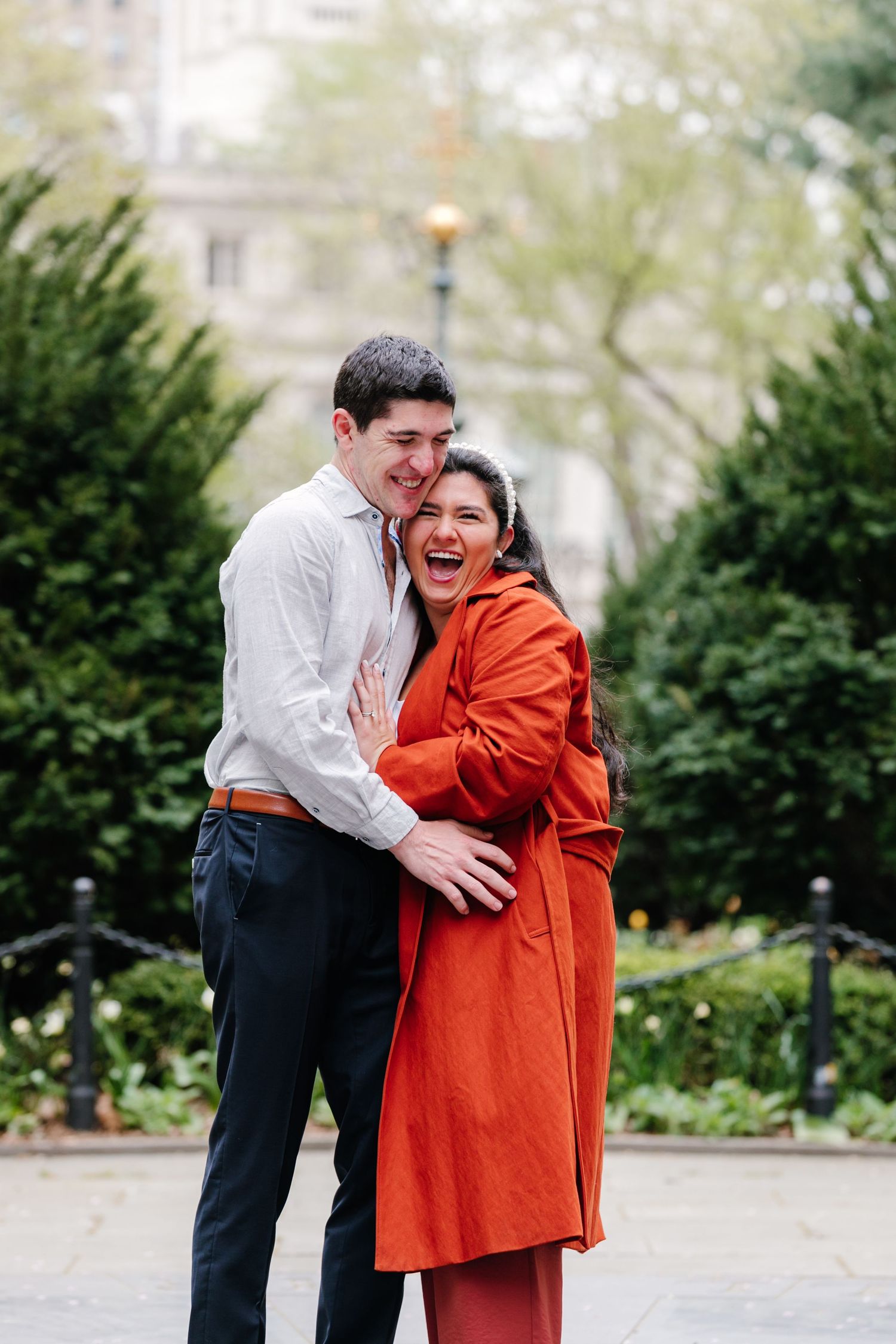 Joyful woman in orange coat laughing while embracing her partner during a spring engagement photo session.