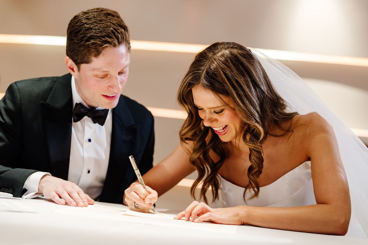 Bride and groom smiling while signing wedding certificate together at elegant ceremony table.
