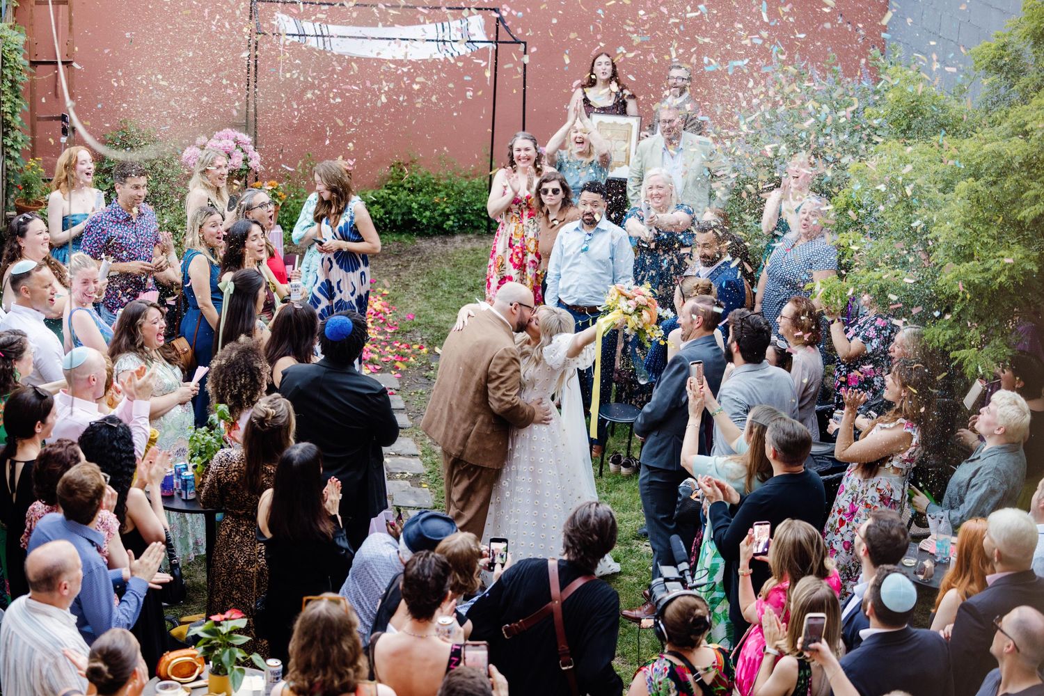 Outdoor wedding ceremony with couple kissing amid confetti, surrounded by joyful guests in a garden setting.