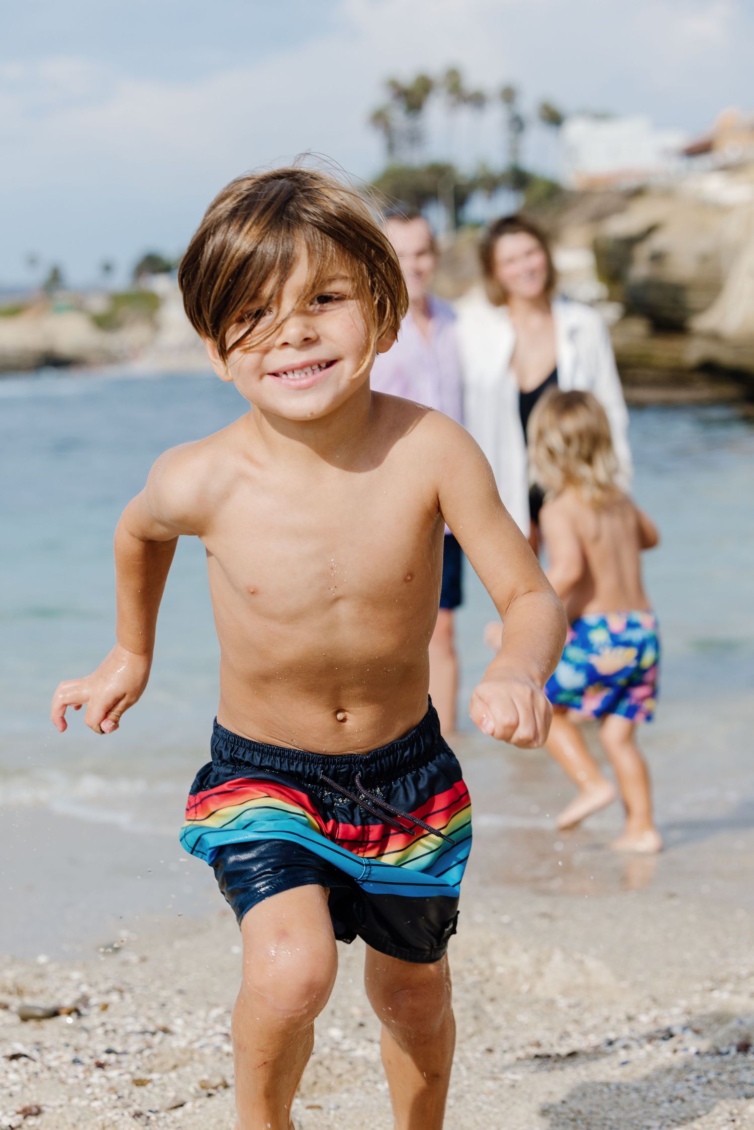 Happy young boy in rainbow swim trunks runs on a sunny beach with his family in the background.