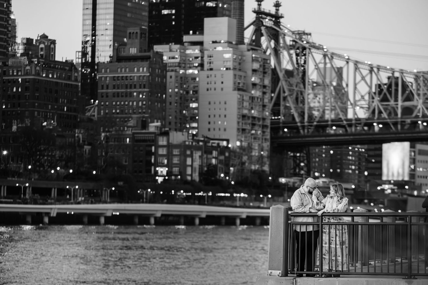 Black and white photo of a couple sharing a romantic moment by a railing with a city bridge and skyline at dusk.