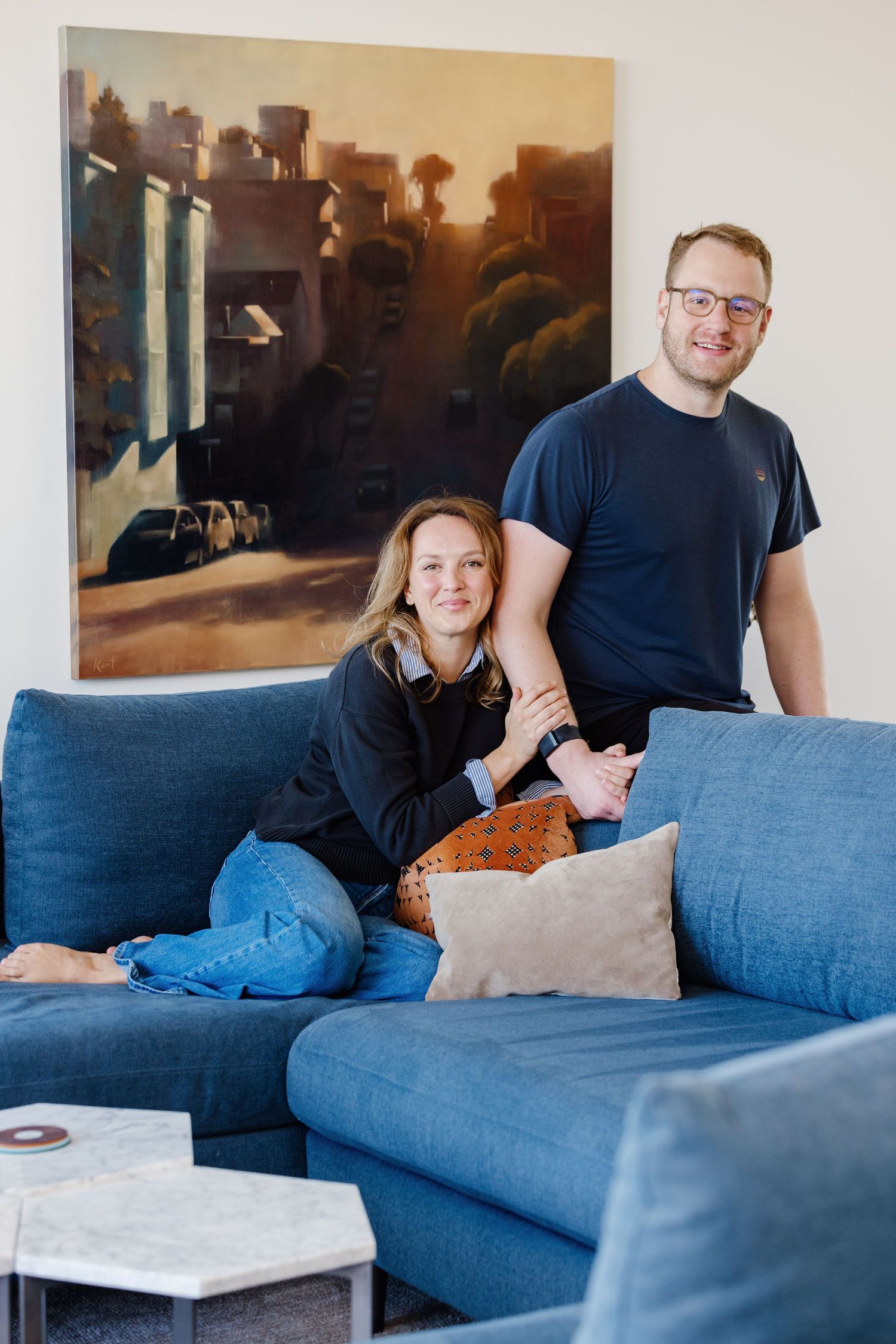 Happy couple posing in a stylish living room with a blue sofa and urban landscape painting behind them.